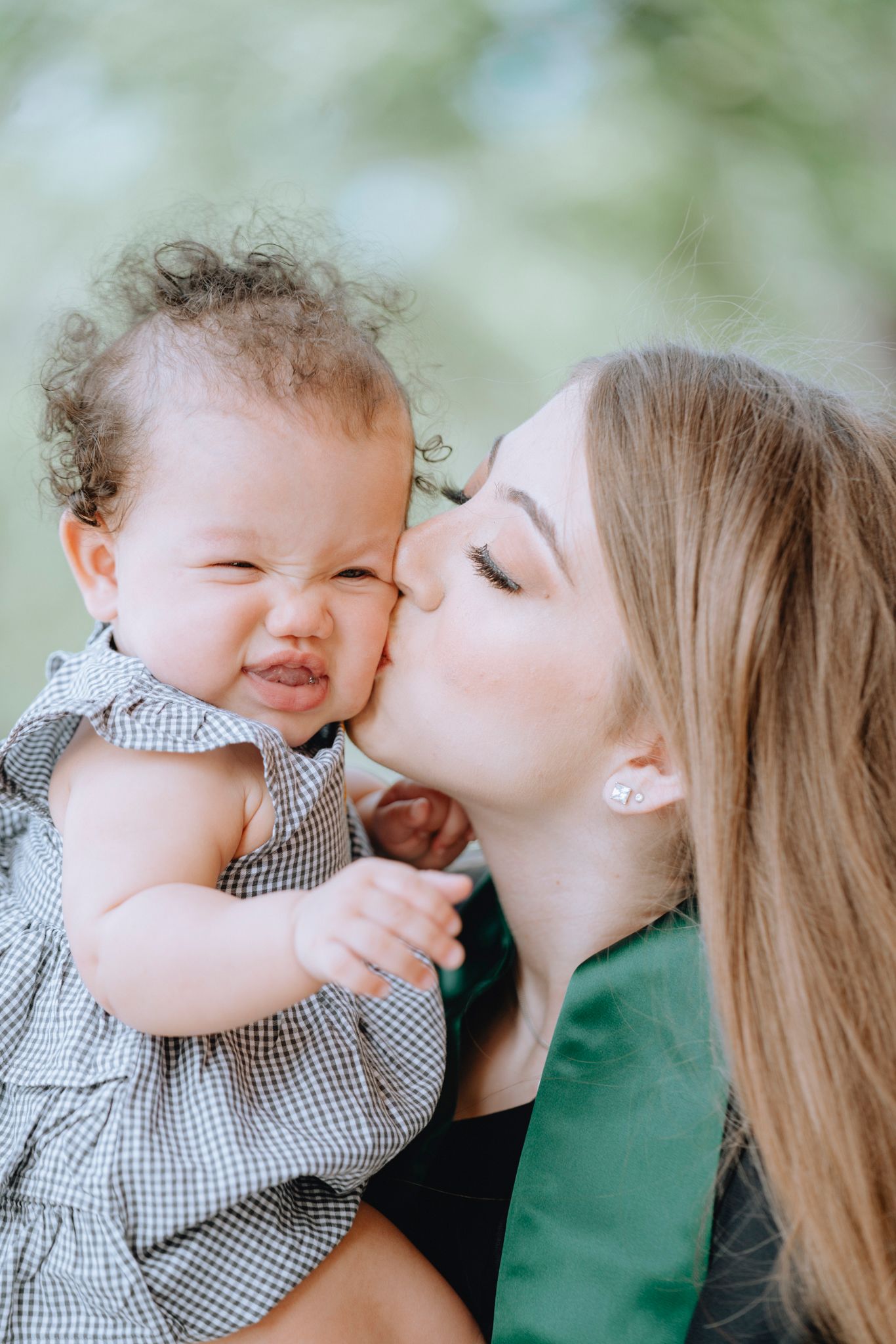 Young woman kissing a curly-haired baby on the cheek outdoors in green setting.