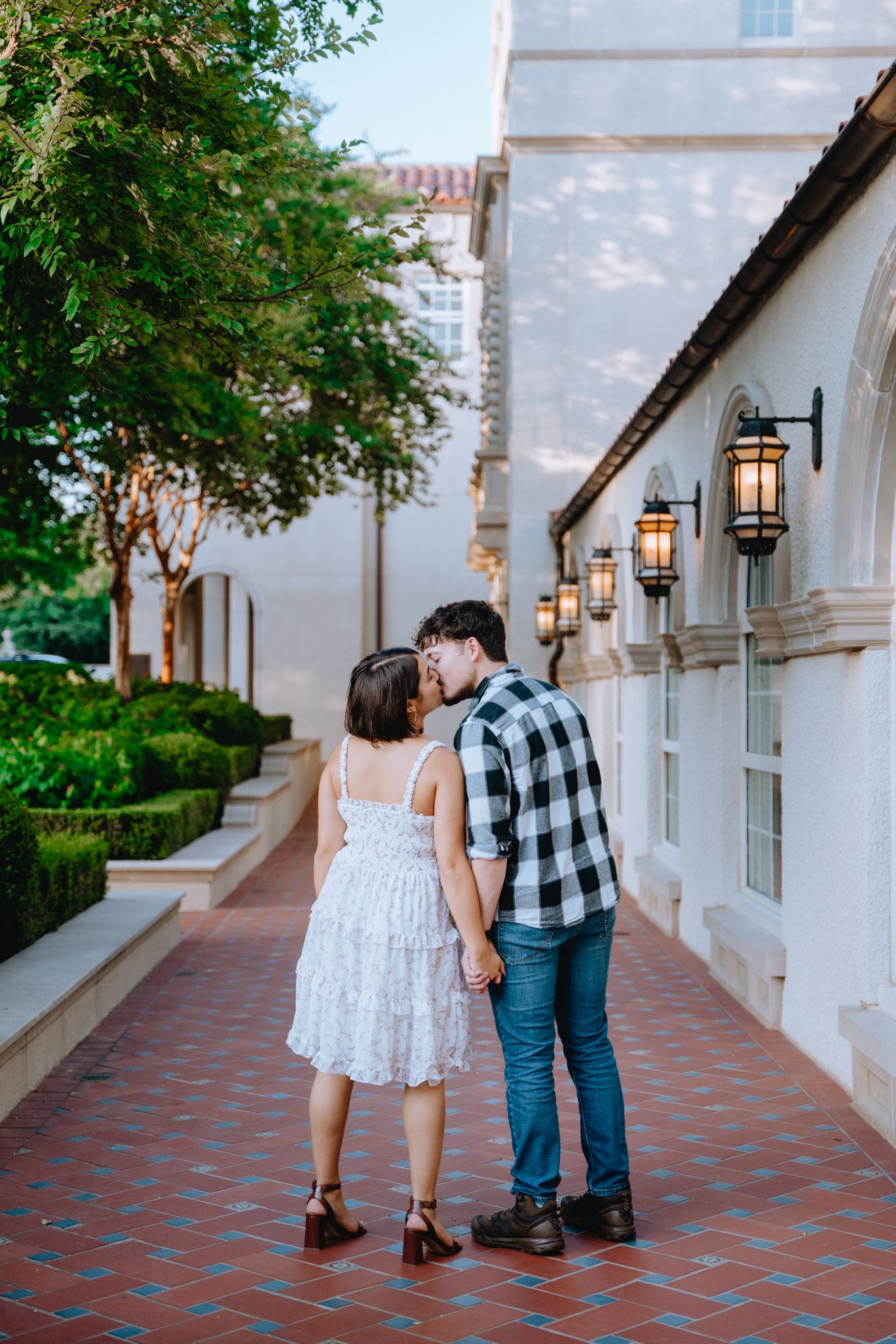 Couple kissing while holding hands on a brick pathway beside a white Spanish-style building.