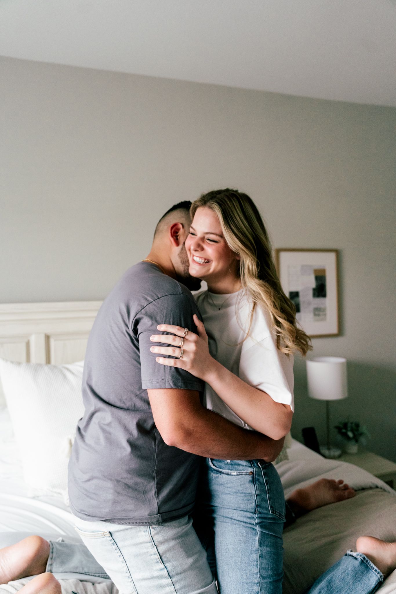 Smiling woman in white top embracing man in gray shirt while sitting on bed in bright bedroom.
