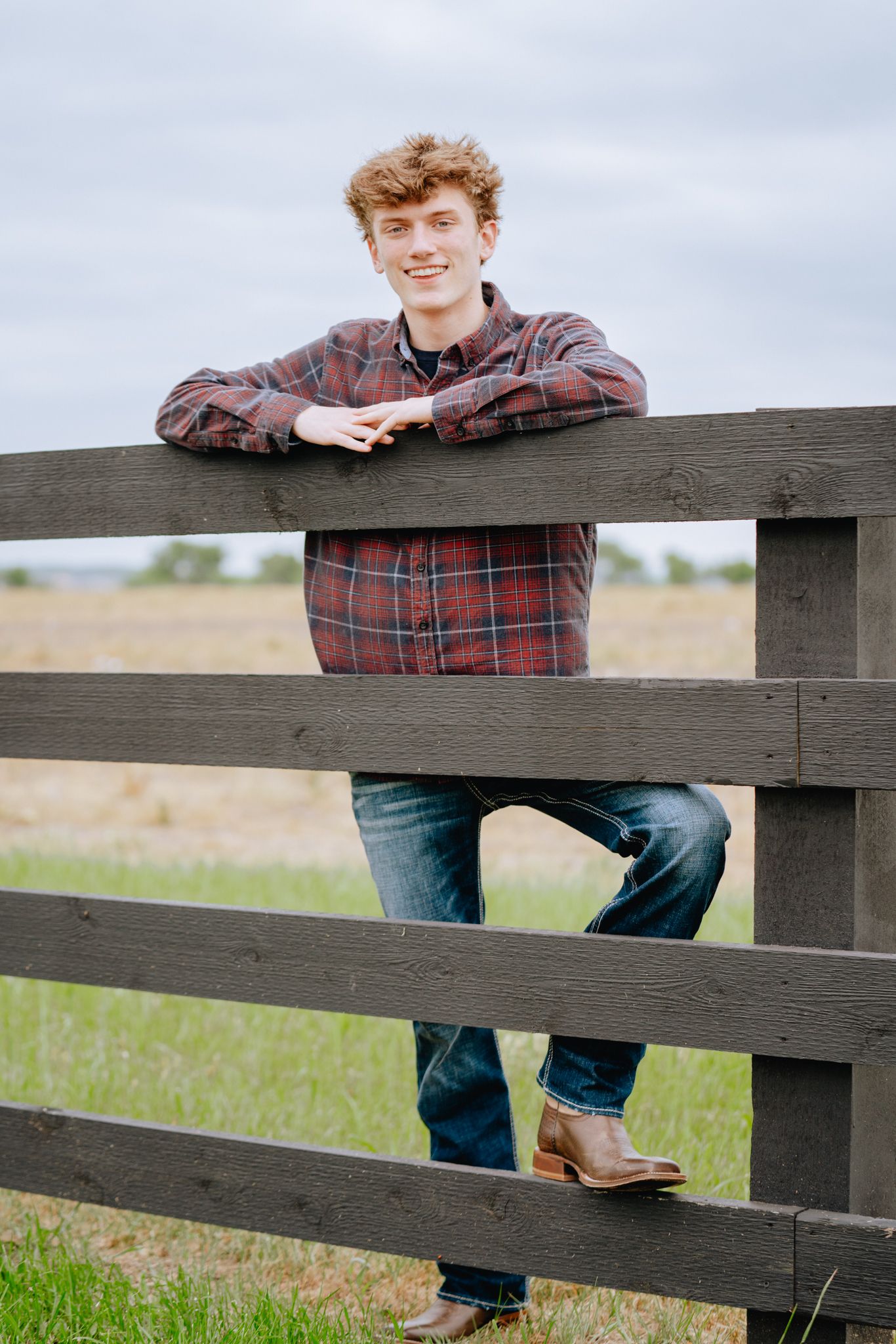 Smiling teenage boy in plaid shirt leaning on rustic wooden fence in a rural field setting.