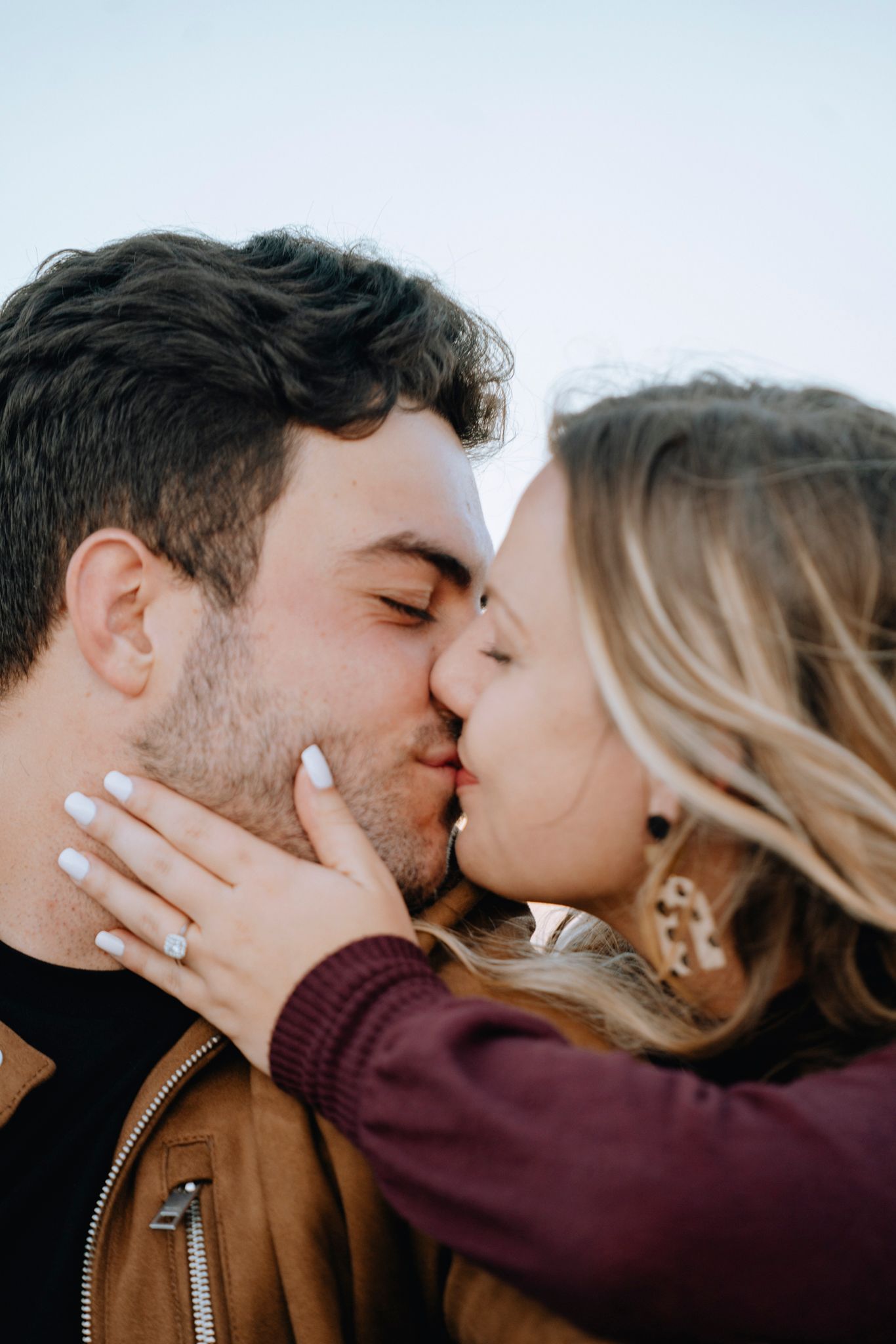 Close-up of engaged couple sharing a kiss, woman displaying diamond engagement ring on mans cheek.