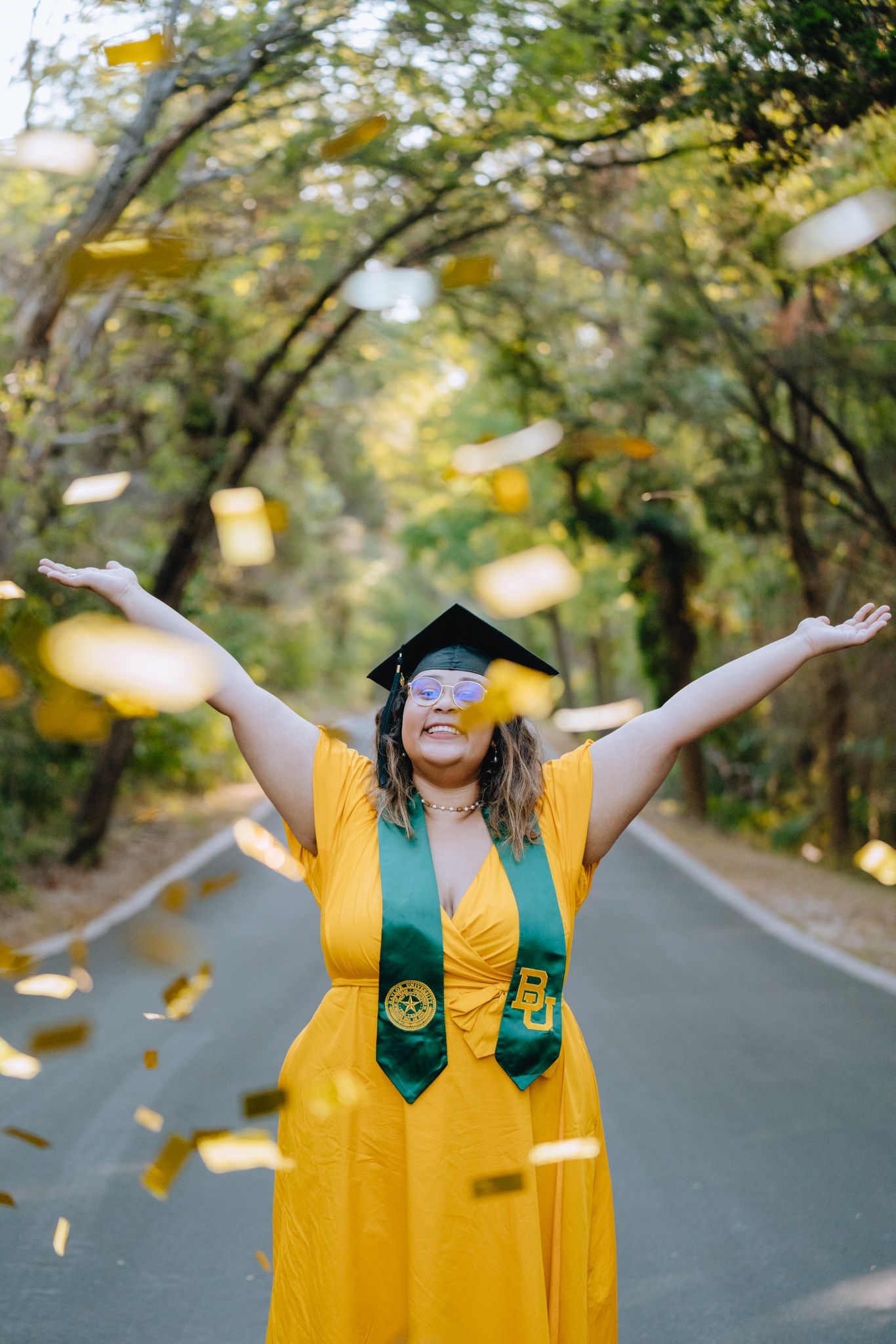 Graduate in yellow dress and cap tossing gold confetti on a tree-lined path.