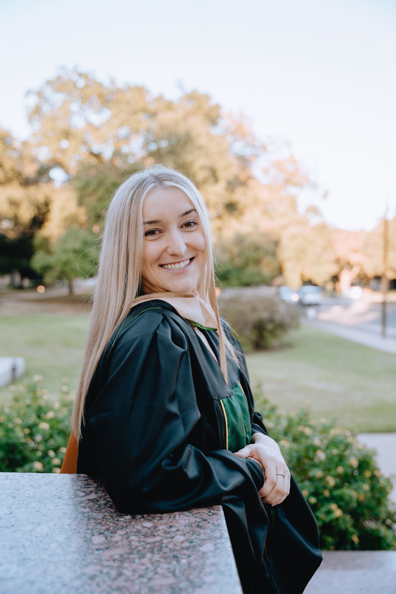 Smiling blonde graduate in green graduation gown posing outdoors on a sunny day with trees in the background.