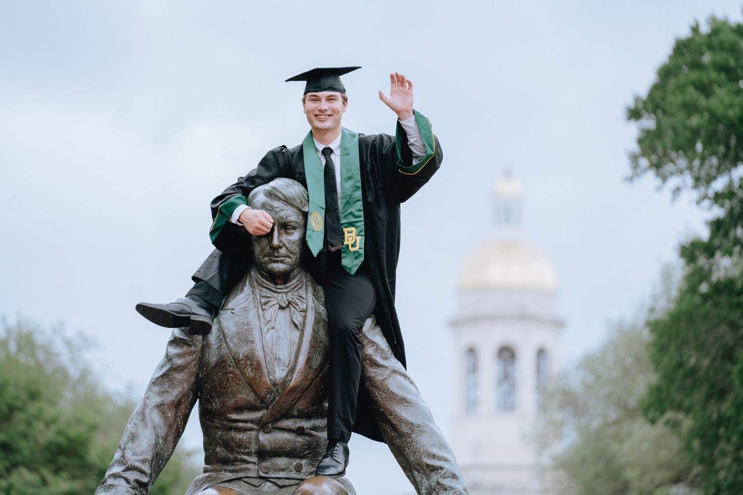 Graduate in cap and gown posing atop a bronze statue with dome building behind.