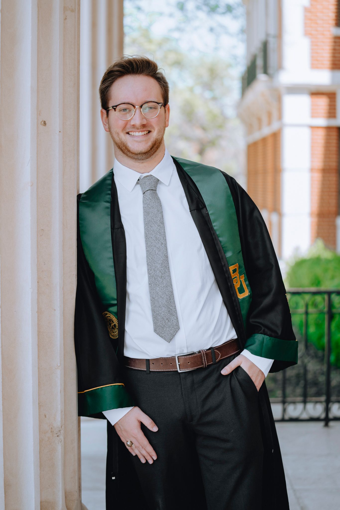 Smiling young man in graduation gown and gray tie posing between columns outdoors.