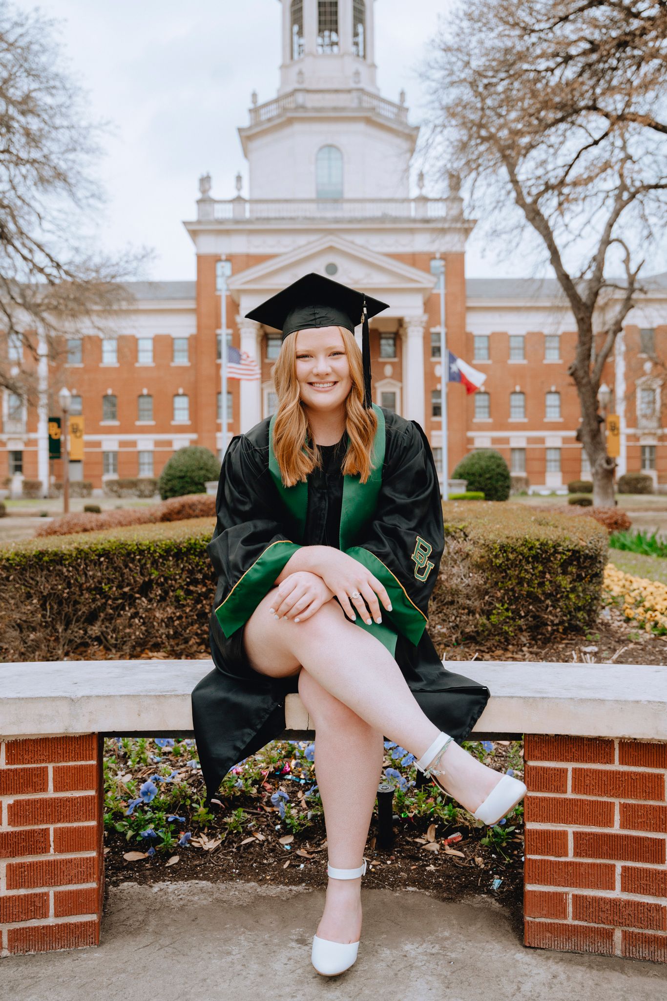 Smiling graduate in black and green cap and gown sitting outside a brick university building.