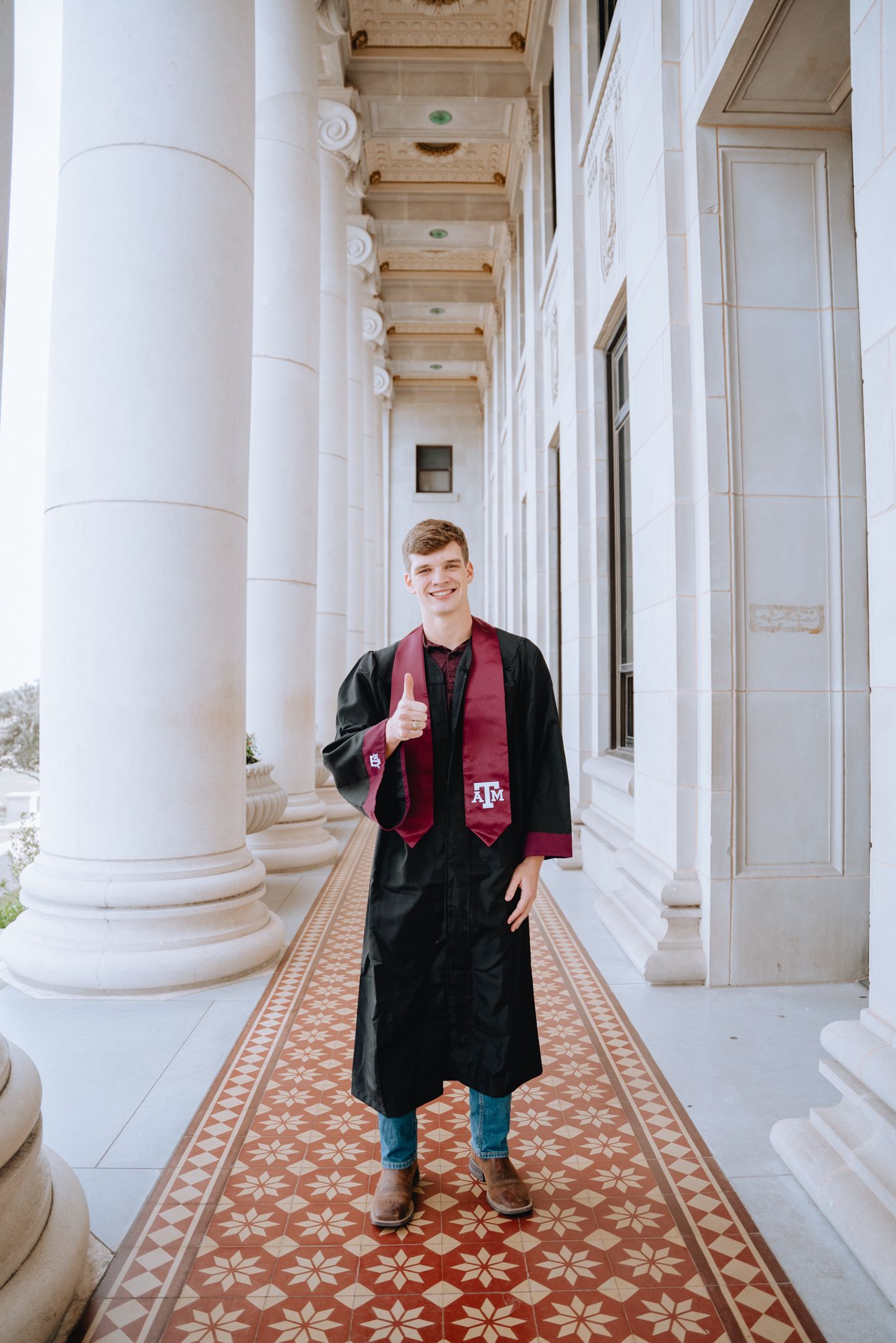 Graduate in black cap and maroon Texas A&M stole giving thumbs up among white columns on campus.