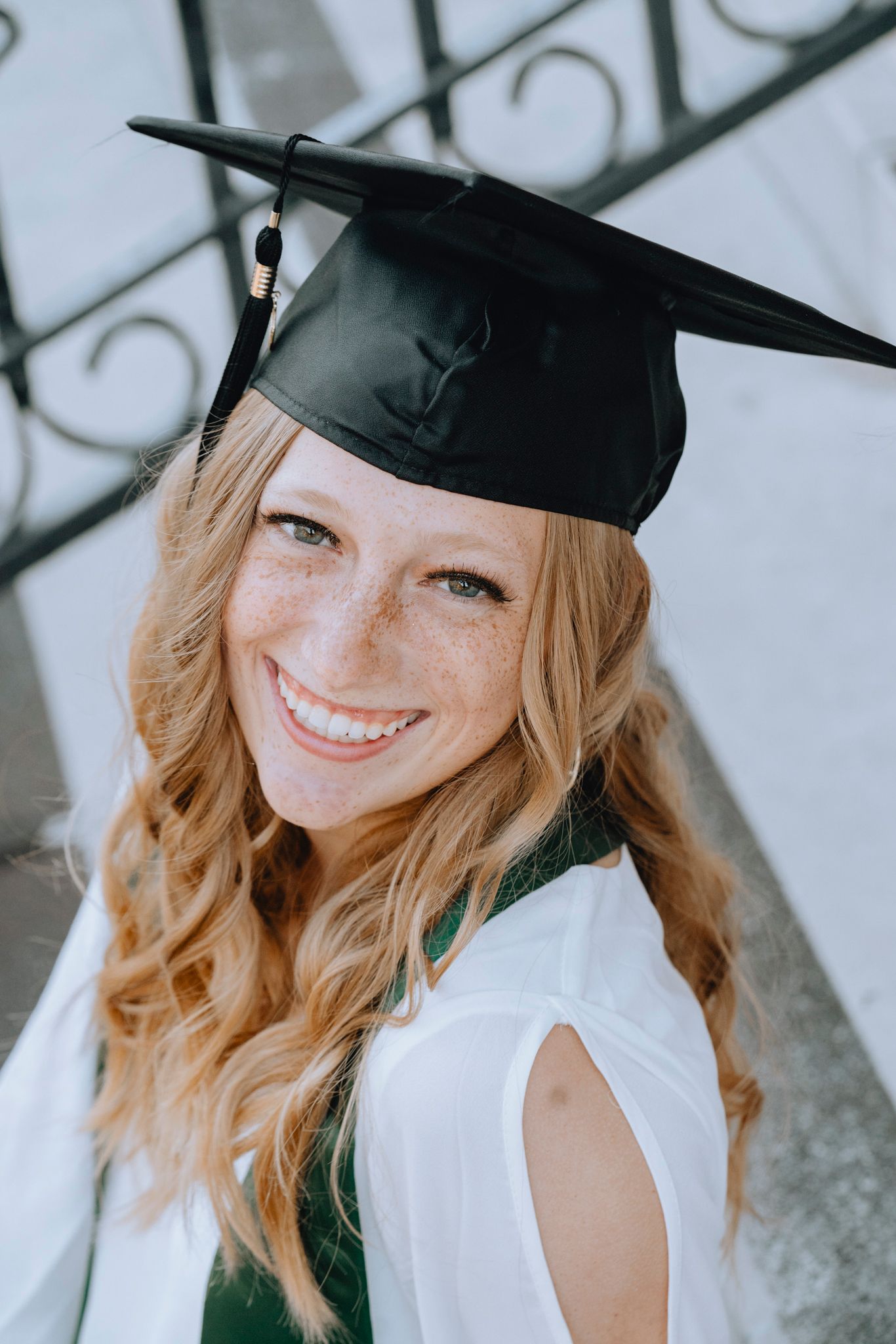 Smiling redhead woman with freckles wearing black graduation cap and green stole outdoors.