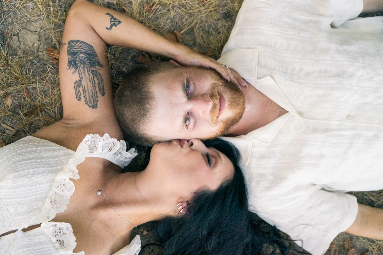 Romantic couple lying on hay, faces close together, man with arm tattoos and woman in white lace dress.