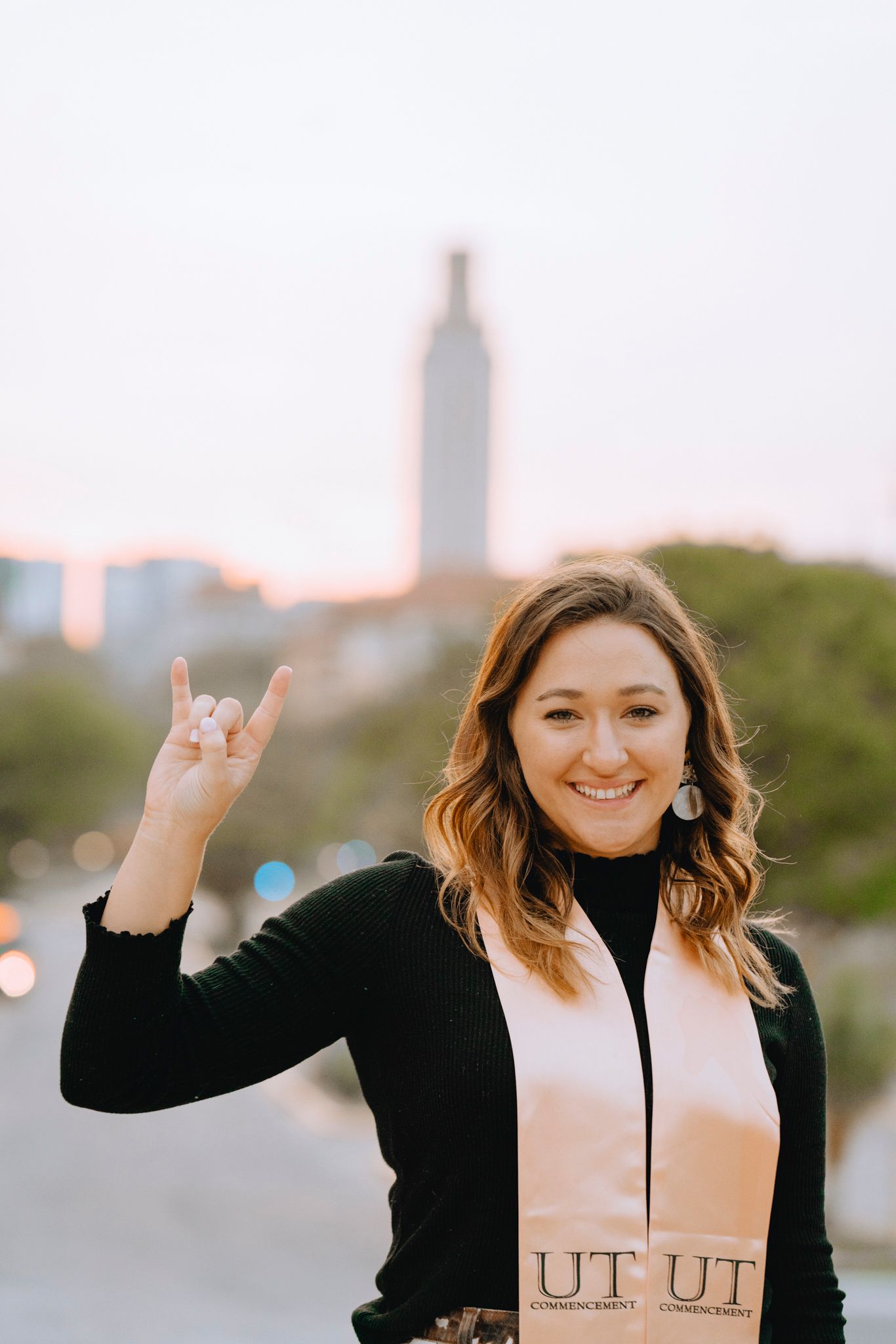 Smiling young woman making hook em horns sign with UT Tower visible in background at sunset.