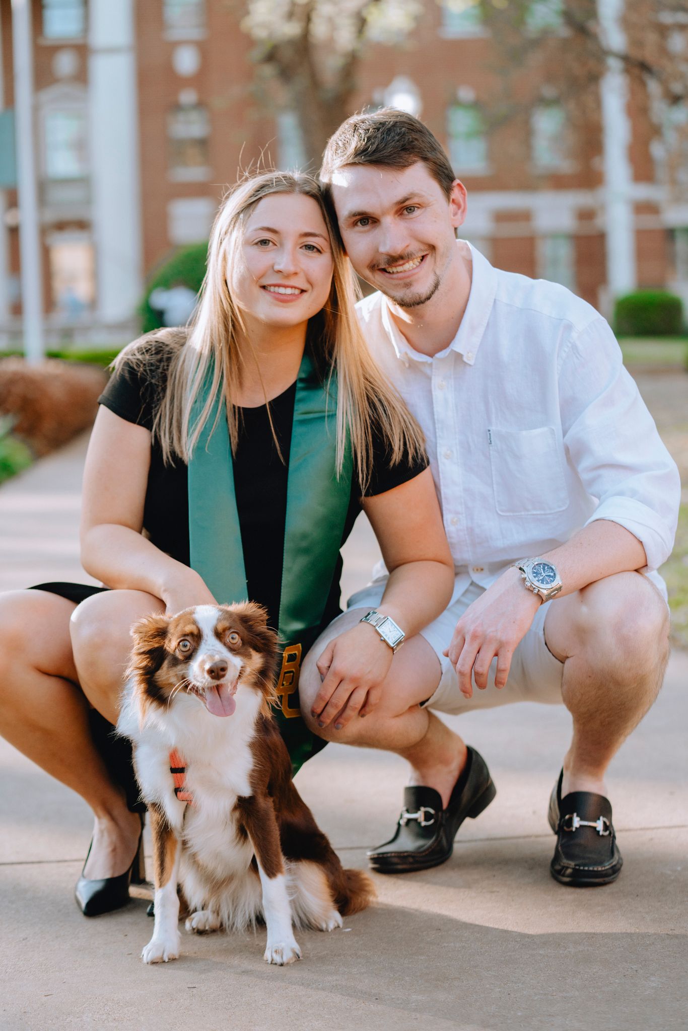 Smiling couple crouching with their brown and white Australian Shepherd dog on campus walkway.
