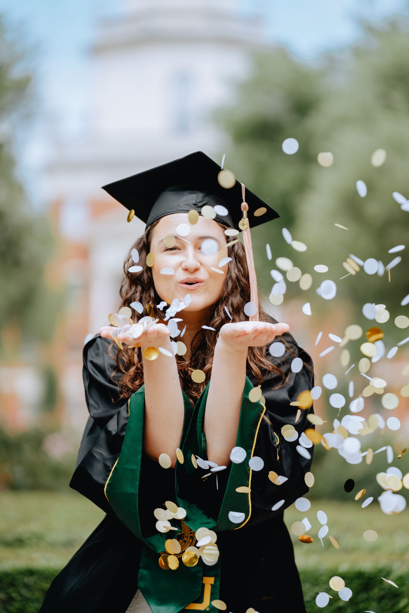 Graduate in cap and gown blowing gold and silver confetti from her hands outdoors.
