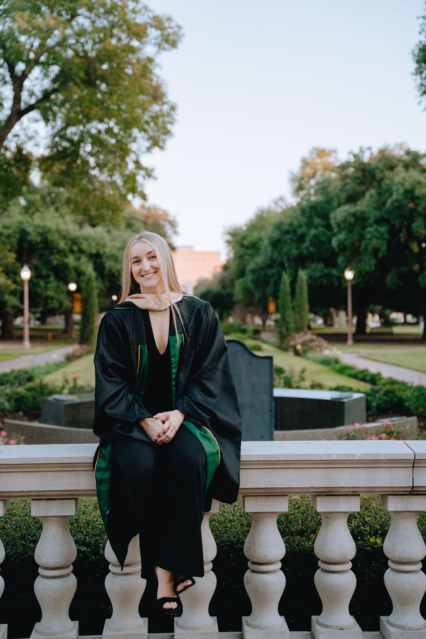 Graduate in black cap and gown sitting on stone balustrade in a garden setting.