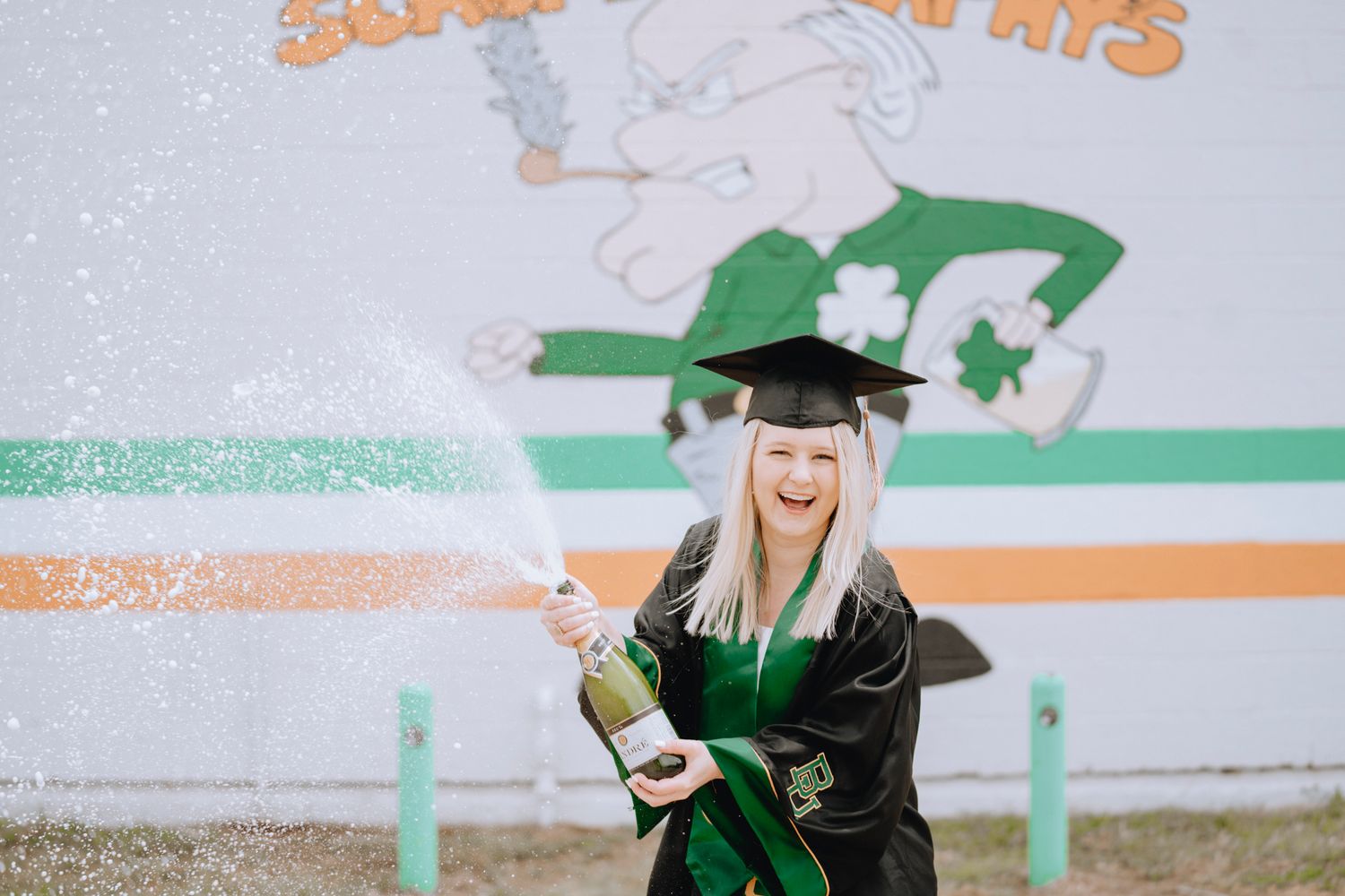 Excited blonde woman in graduation gown popping champagne bottle outdoors near colorful mural.