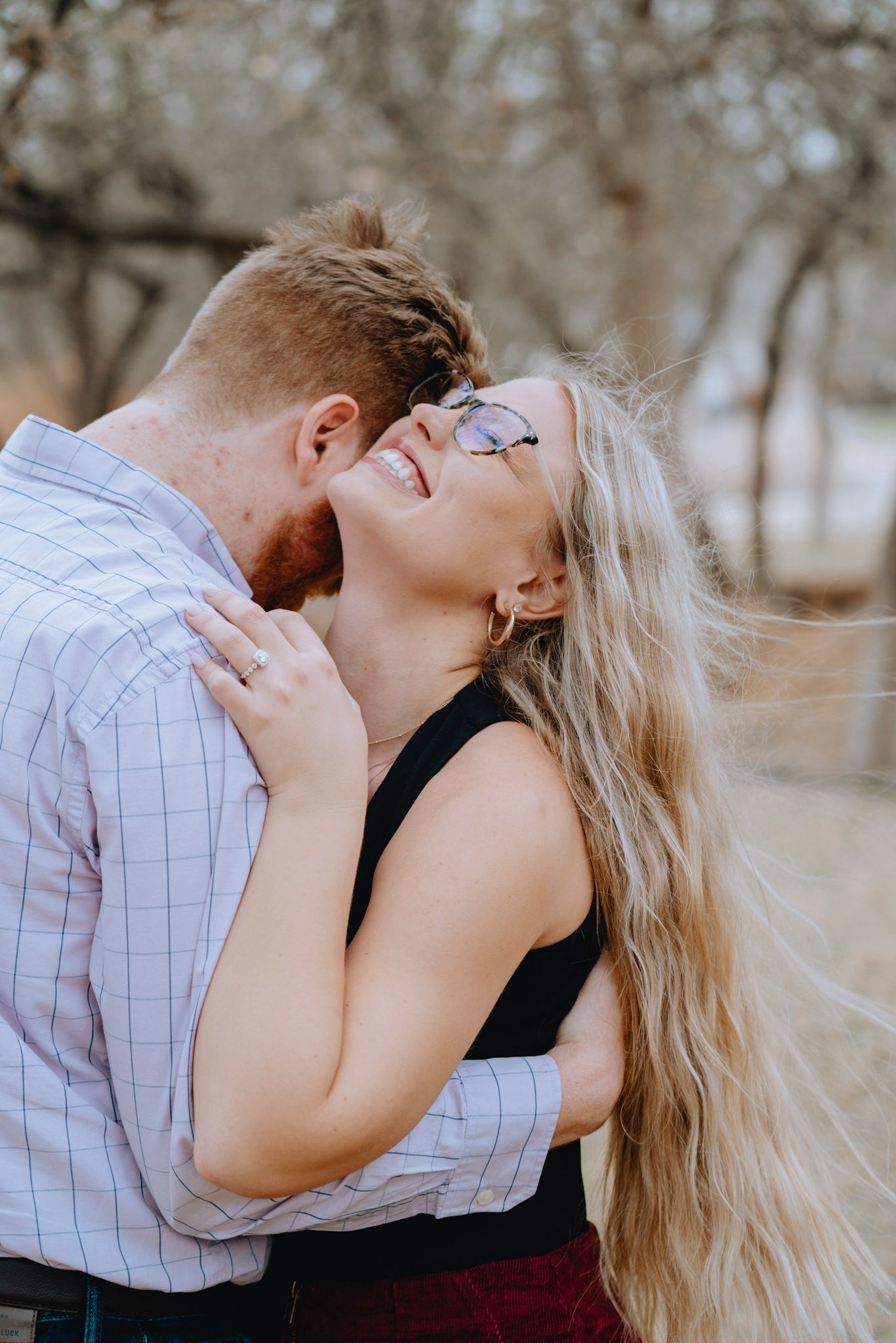 Couple embracing outdoors, woman laughing joyfully as man kisses her neck among bare winter trees.