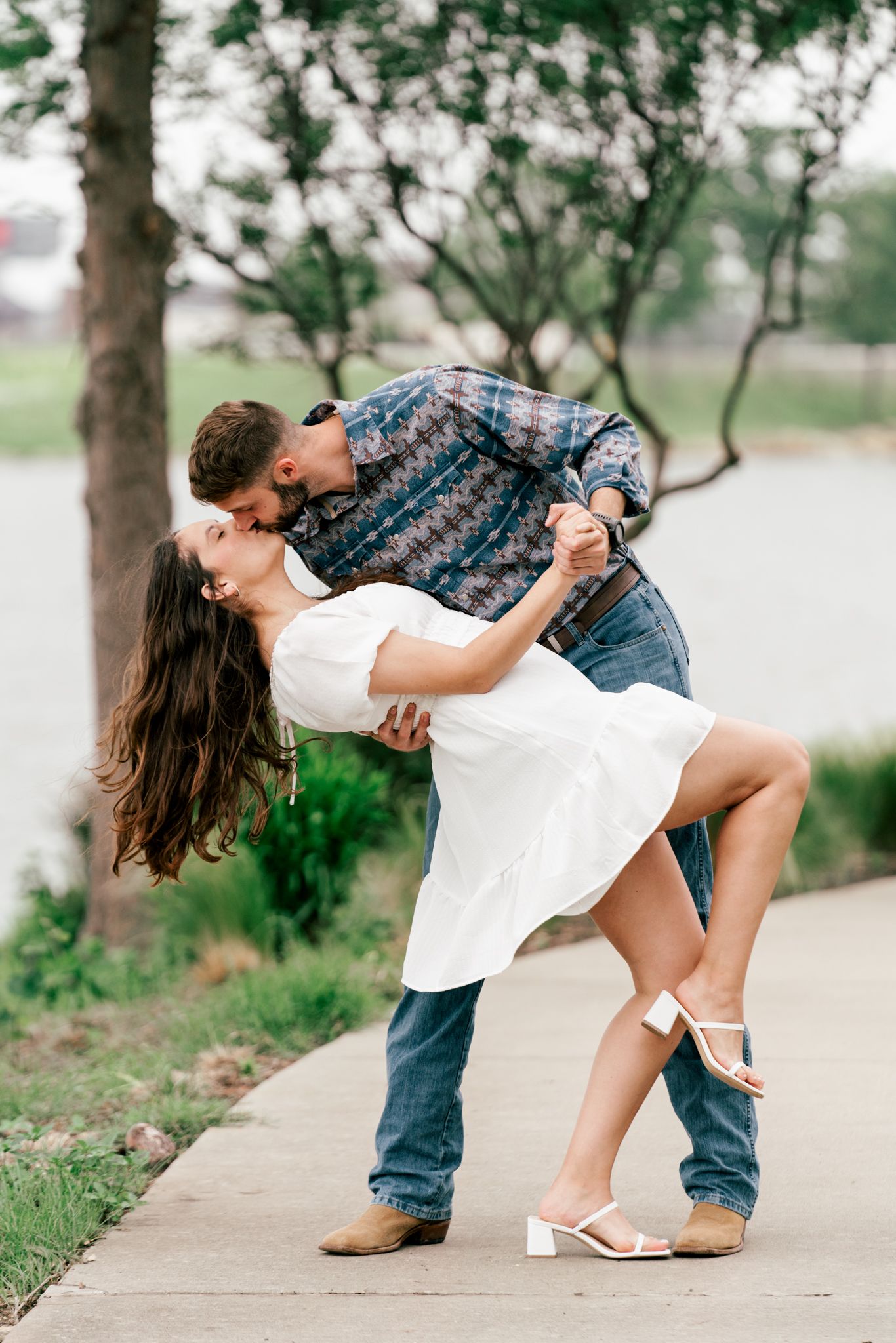Couple sharing romantic dip kiss on pathway near scenic waterfront.