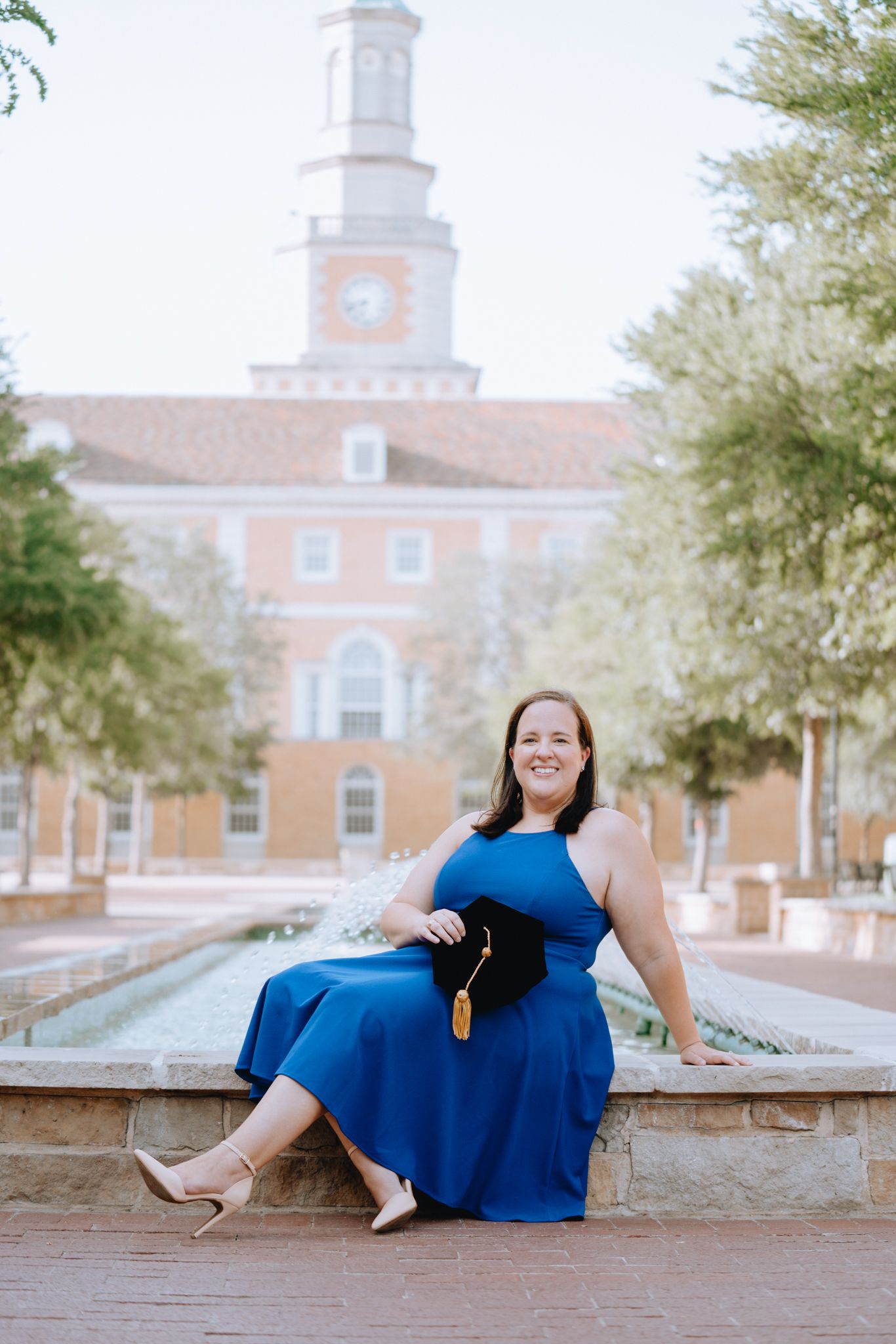 Woman in blue dress sitting by fountain holding graduation cap with university clock tower in background.