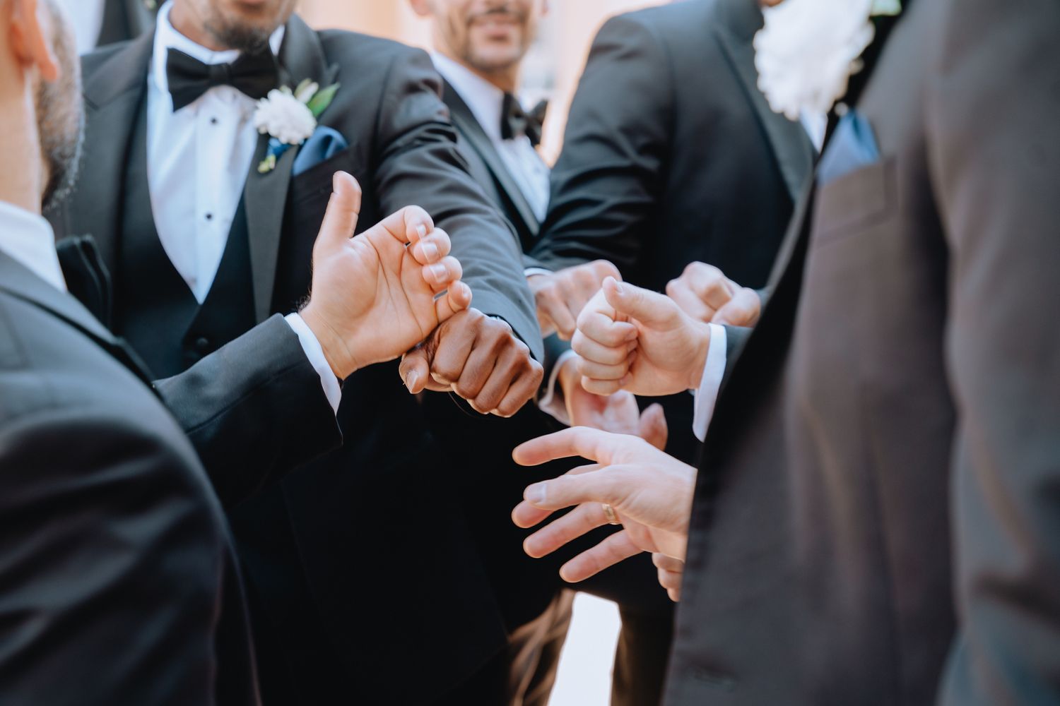 Groomsmen in black tuxedos joining hands together in a celebratory gesture at a wedding ceremony.