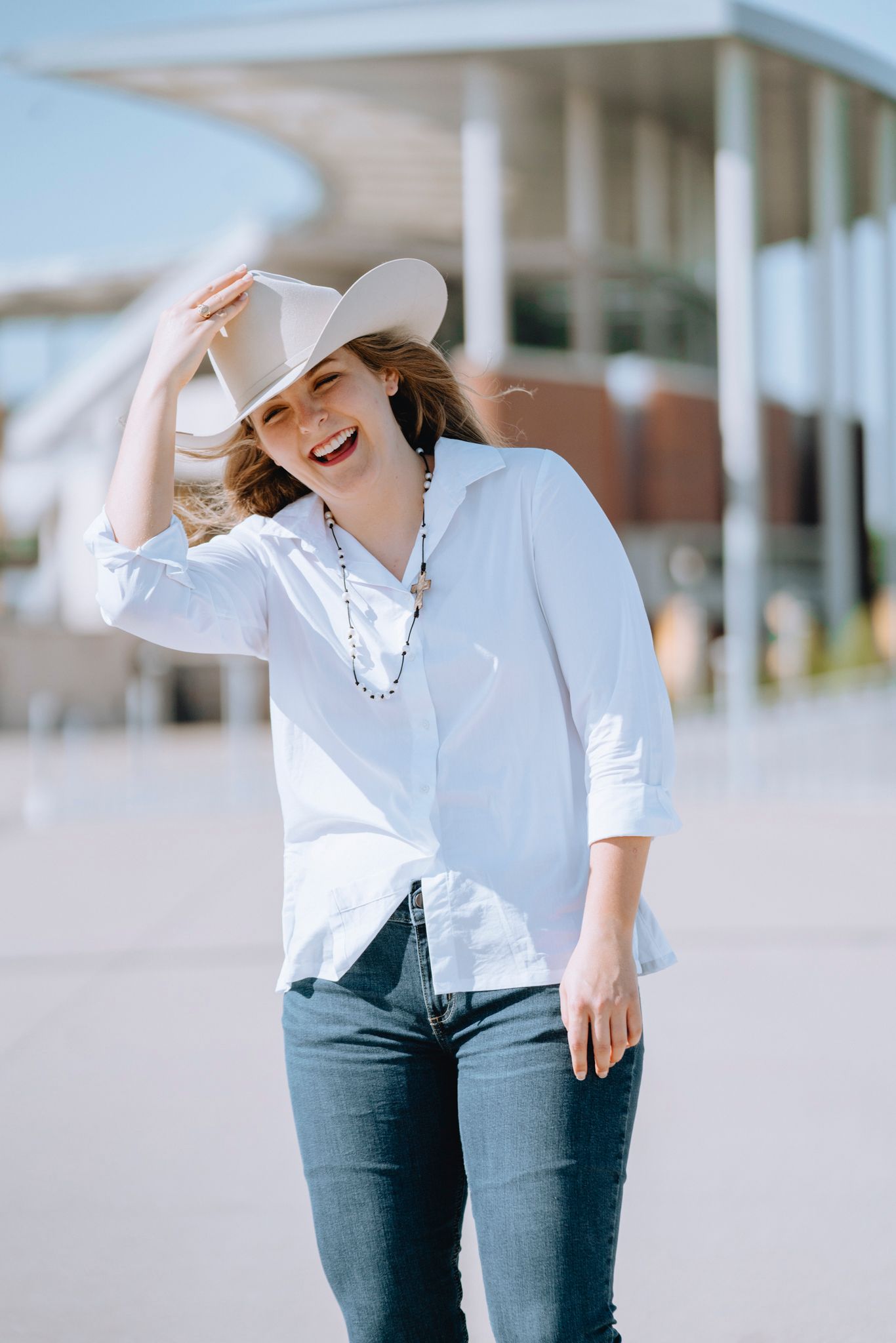 Woman in white shirt and cowboy hat laughing outdoors on a sunny day.