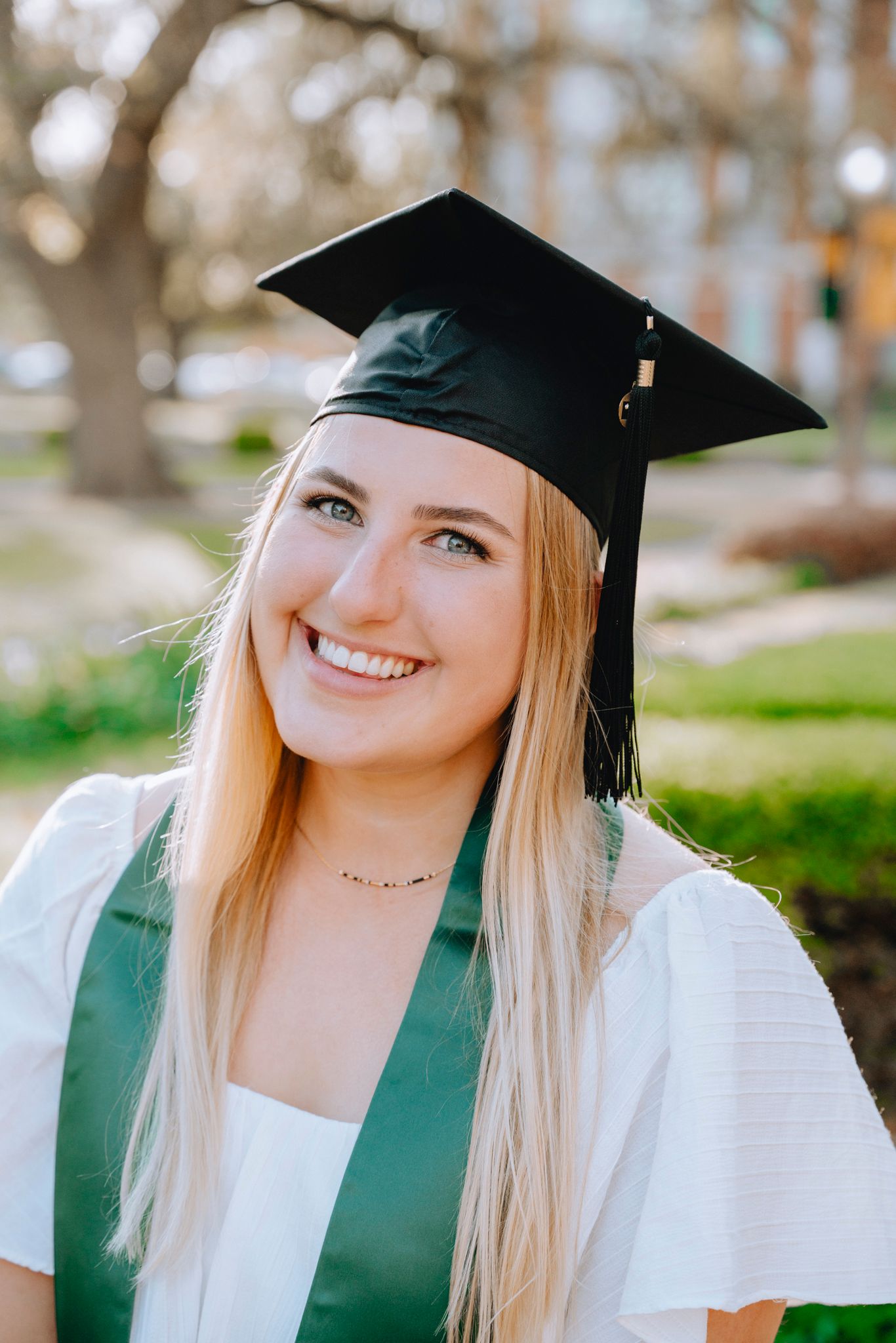 Smiling blonde woman in black graduation cap and green BU stole poses outdoors for graduation photos.