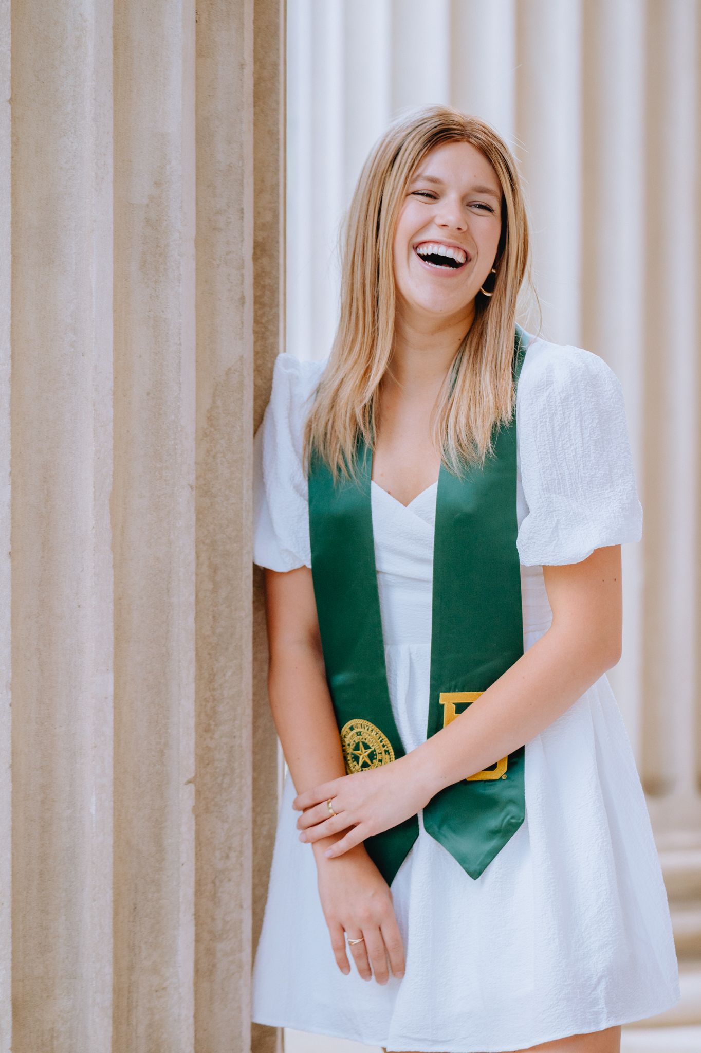 Laughing young woman in white dress with green graduation stole posing between stone columns.