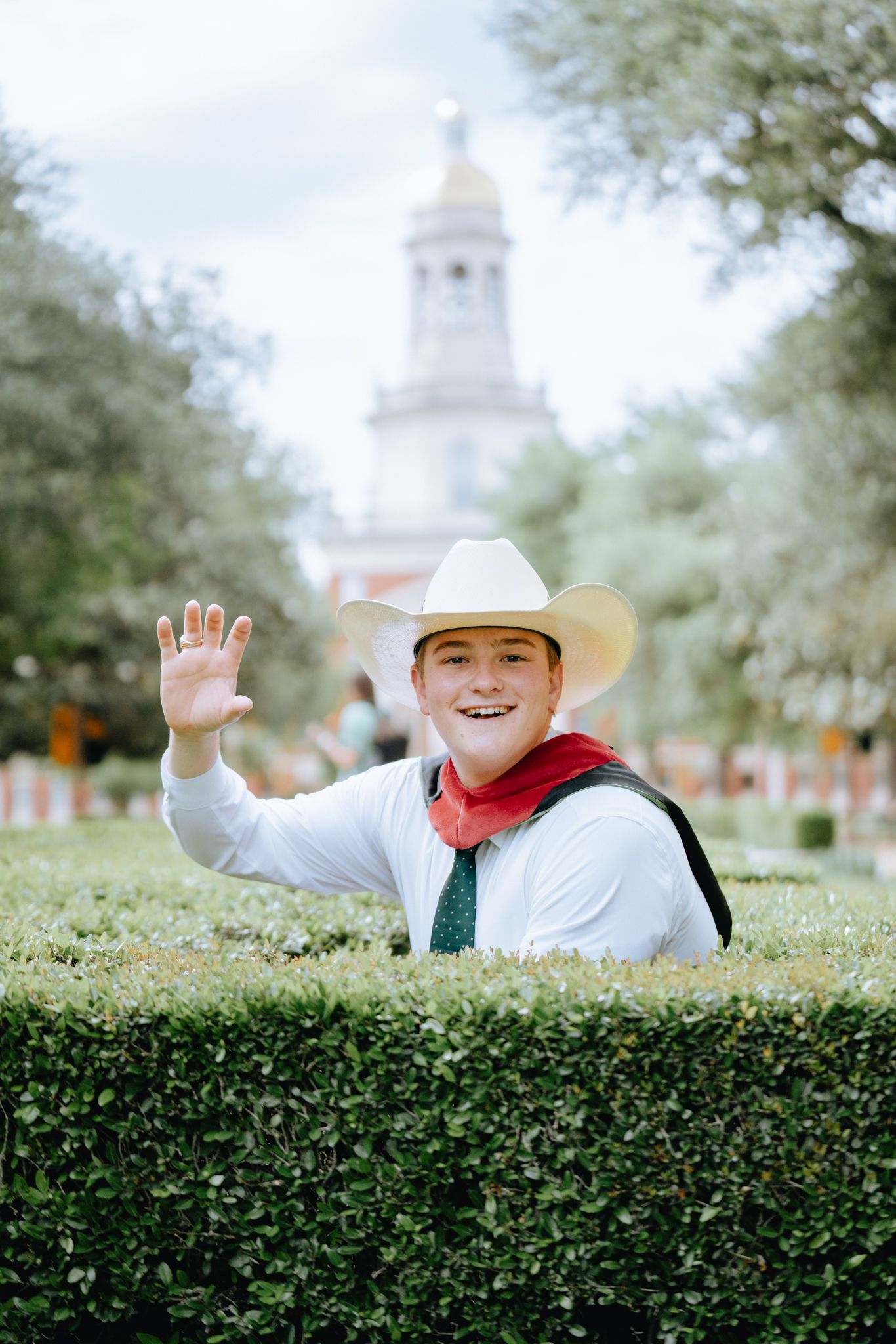 Smiling young man in cowboy hat waving beside hedge with university building behind.