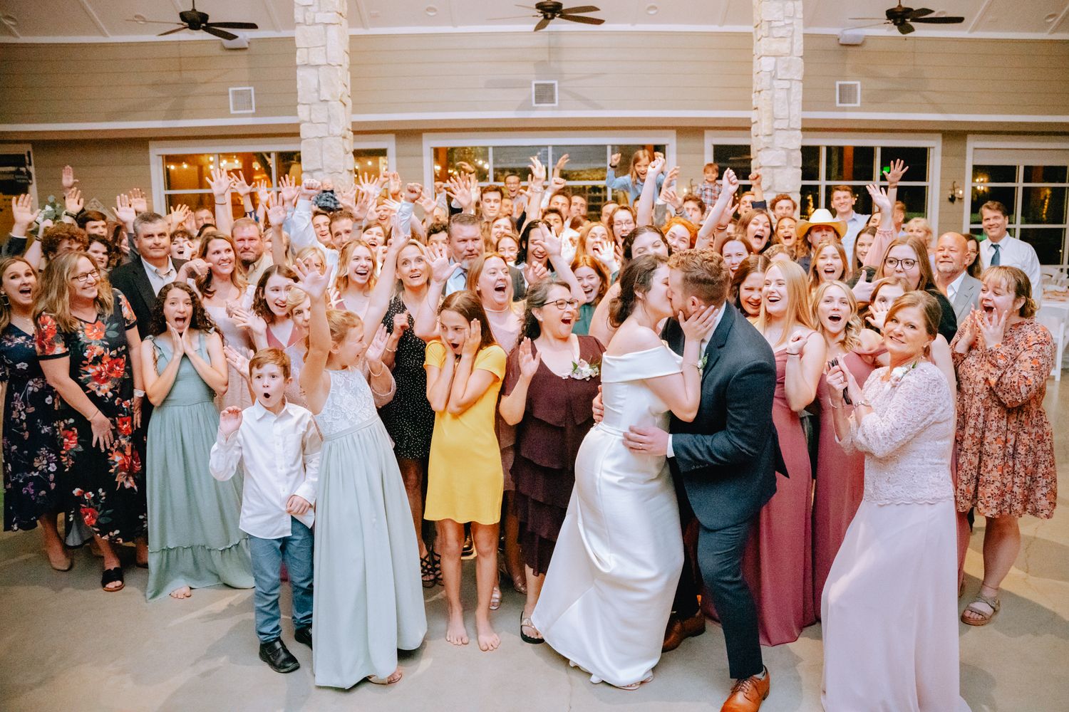Joyful wedding reception group photo with bride, groom, and cheering guests.
