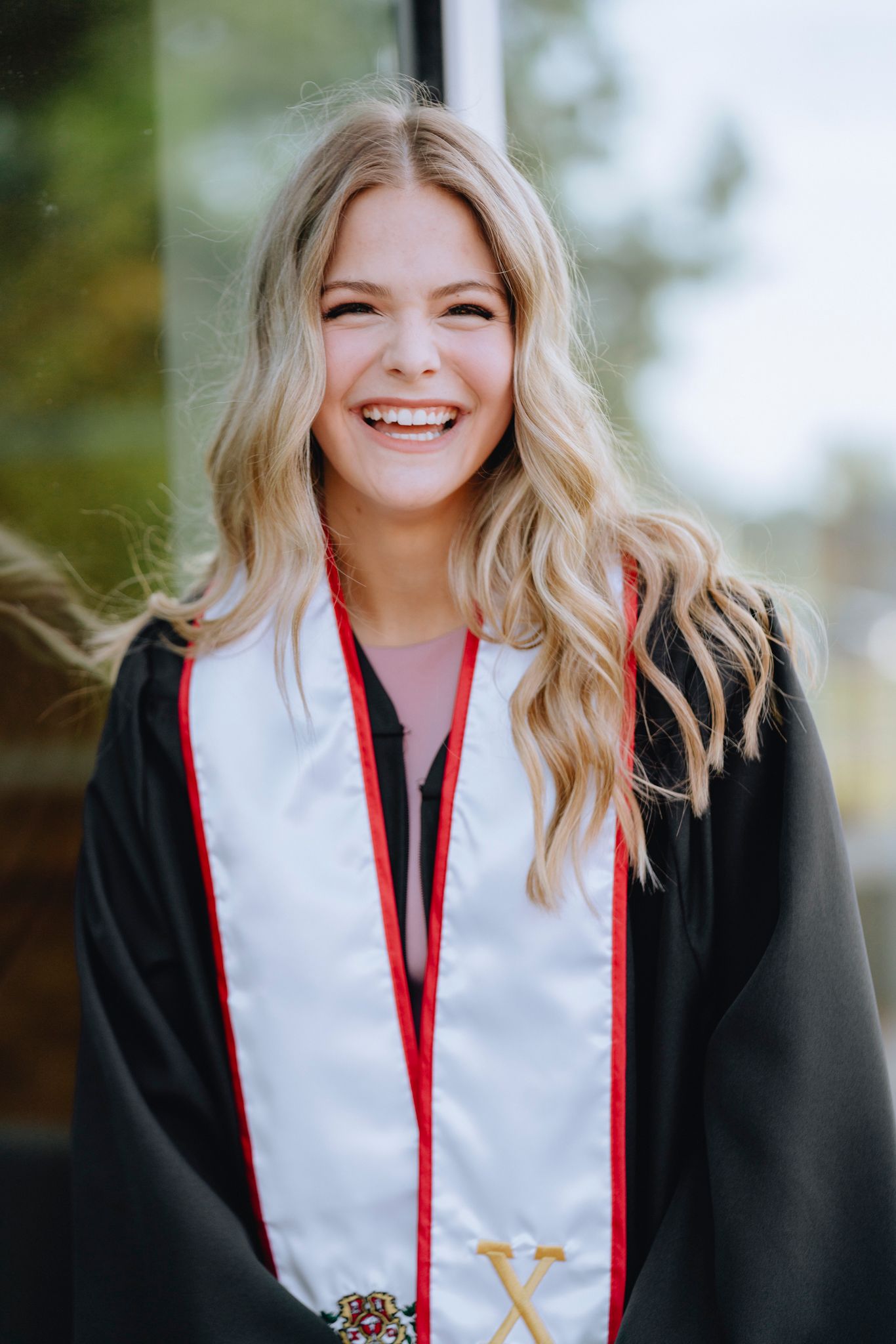 Smiling young blonde woman wearing black graduation gown with white and red stole outdoors.