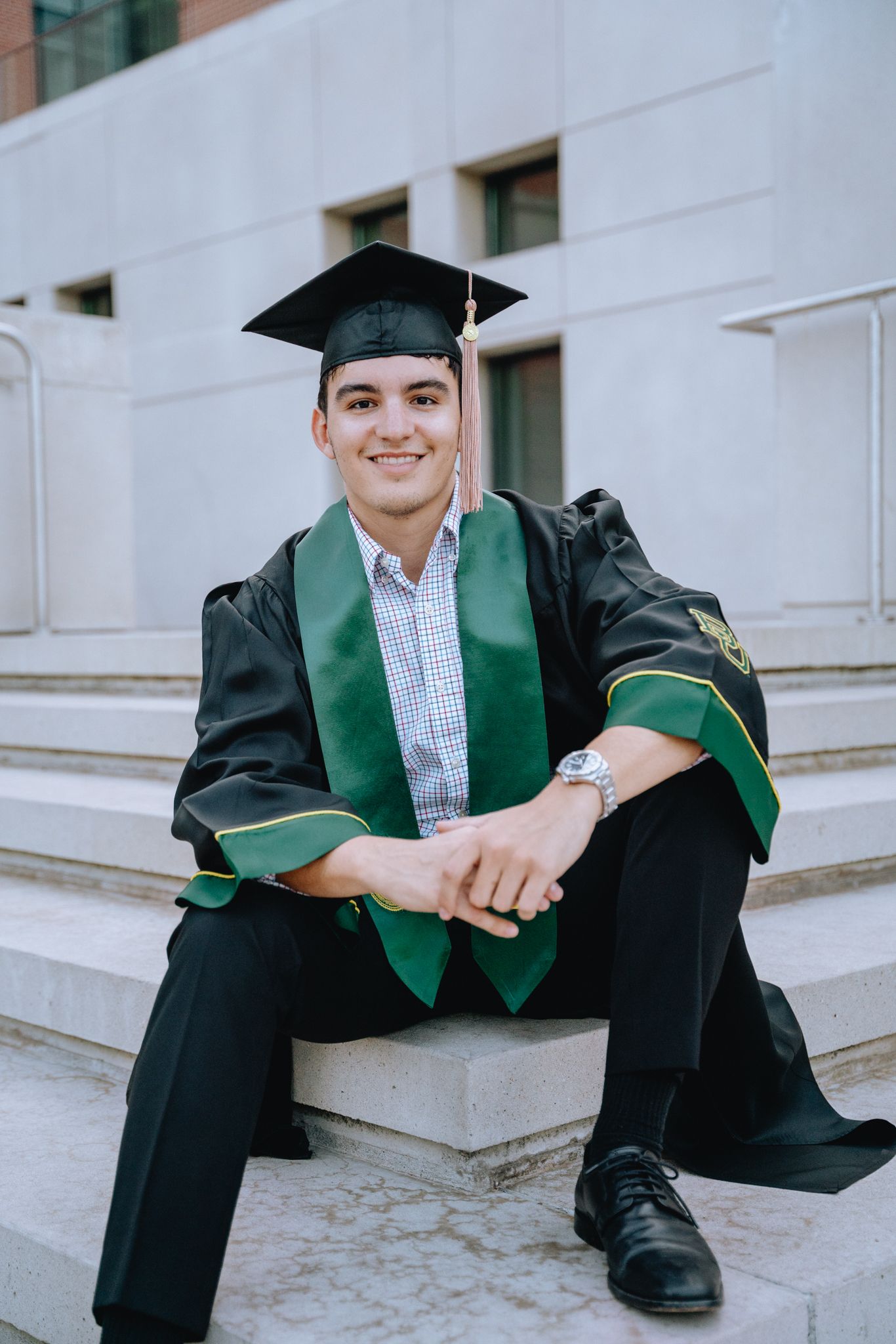 Male graduate in black cap and gown with green stole sits on steps of campus building.