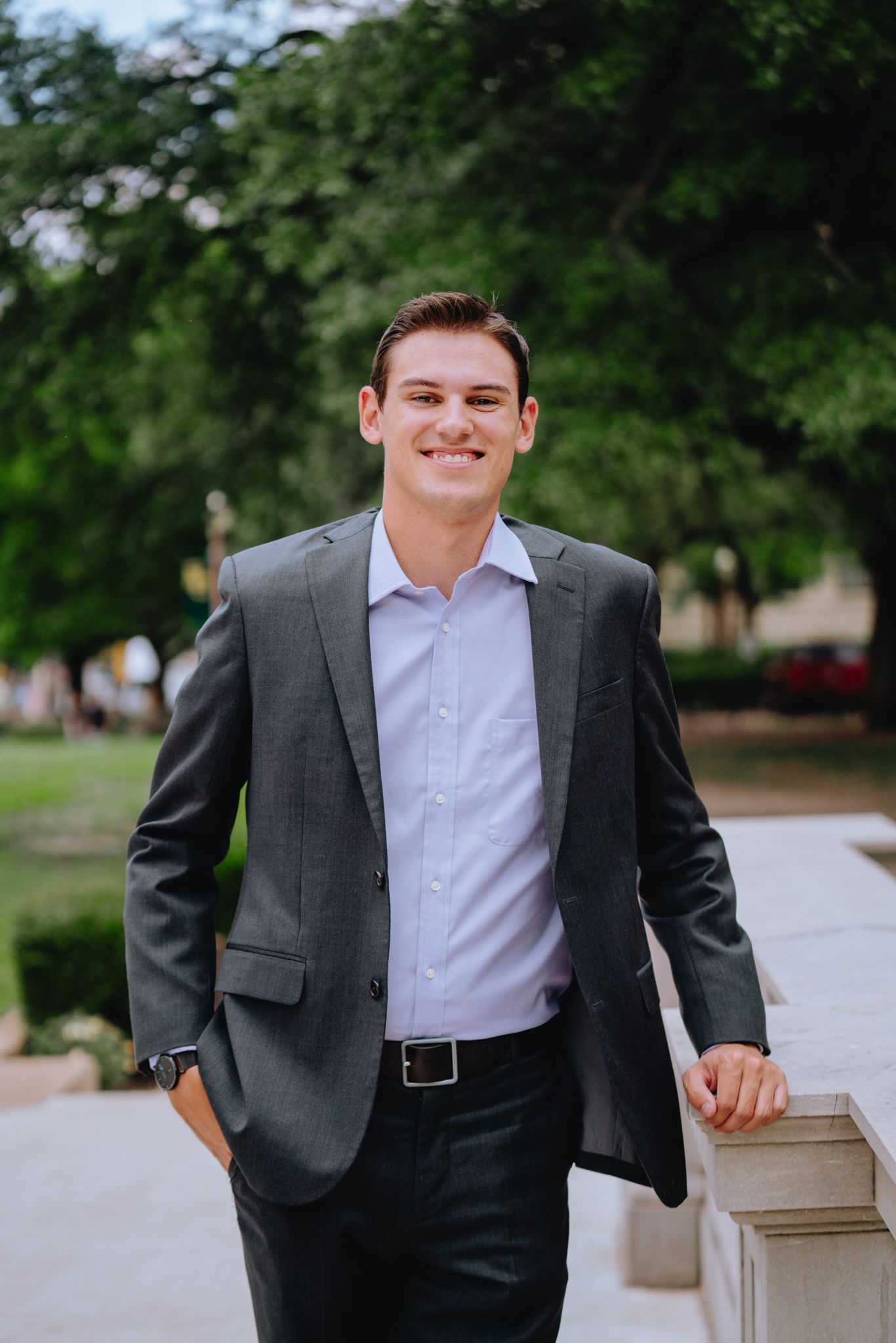Young man in gray suit smiling while leaning against stone wall with green trees in background.