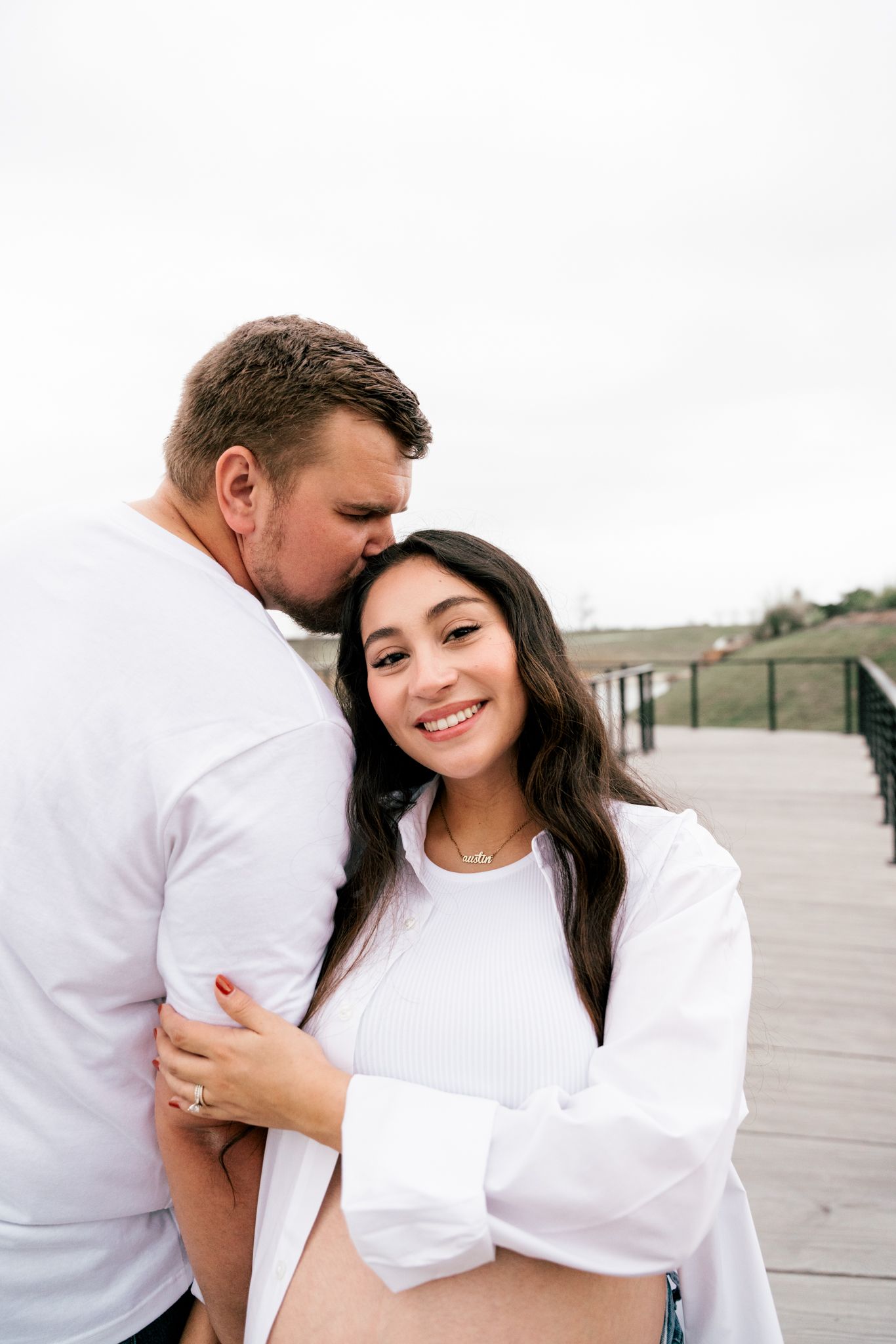 Couple embracing on boardwalk, man kissing woman's head while she smiles, both wearing white outfits.