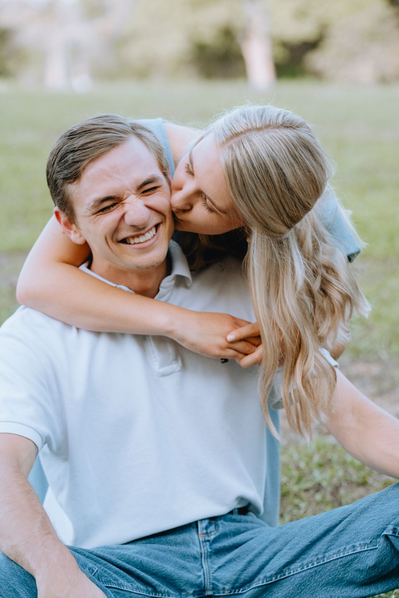 Blonde woman kissing smiling man on cheek in green park.