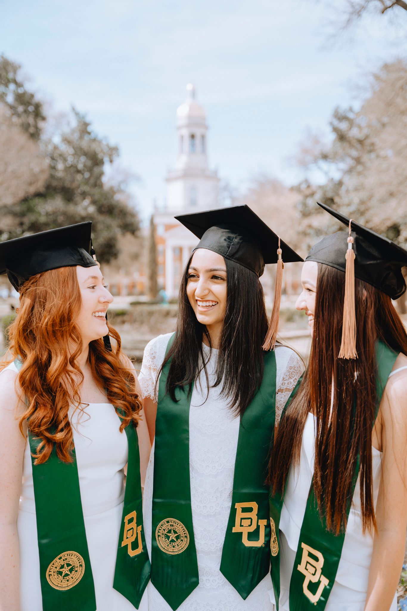 Three Baylor University graduates in green BU stoles and black caps smile together on campus.