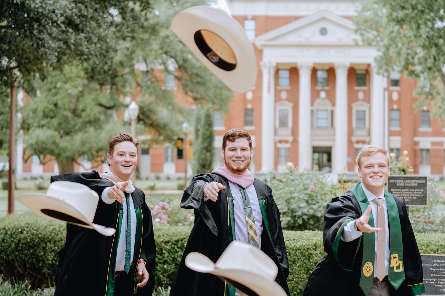 Three Baylor University graduates in black robes toss cowboy hats in front of campus building.