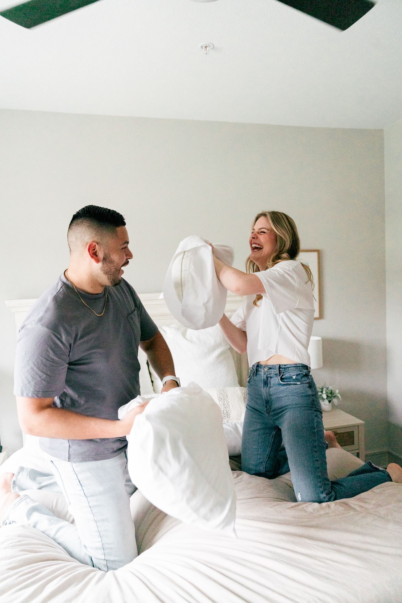 Couple laughing during playful pillow fight on bed in bright bedroom.