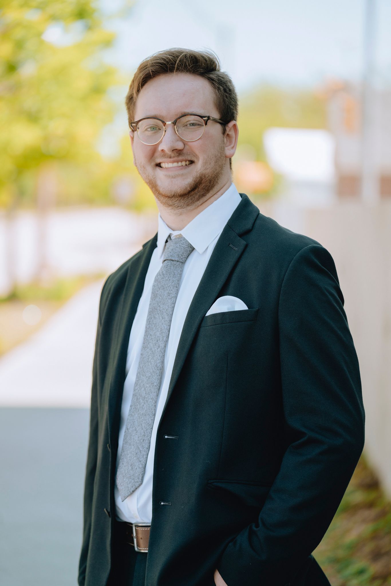 Young man in dark suit and gray tie smiling outdoors for professional portrait photo.