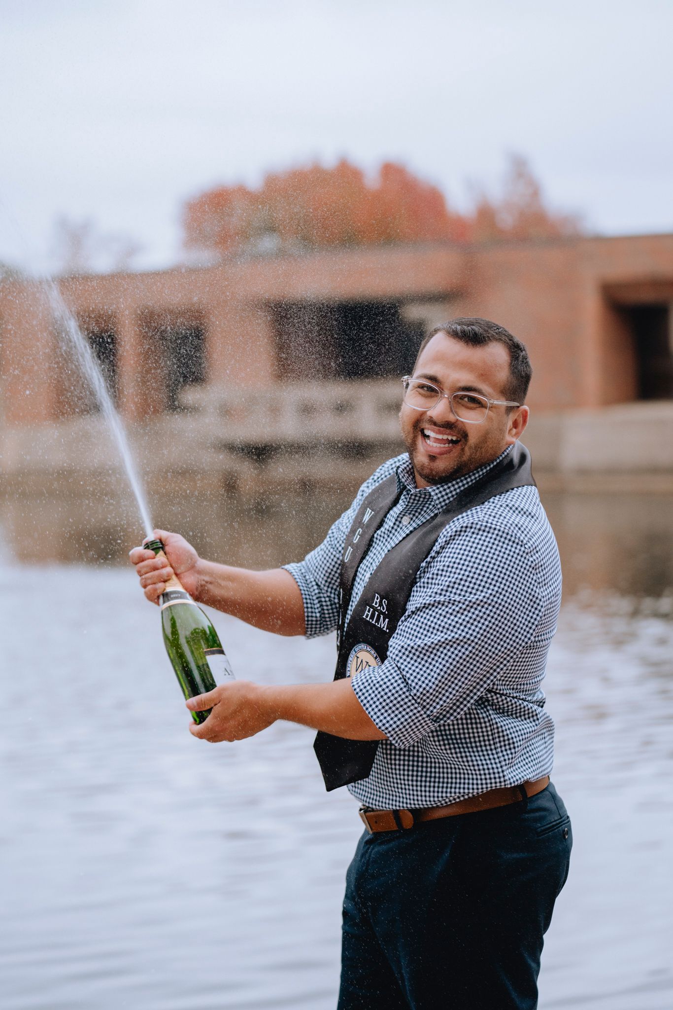 Man celebrating with champagne bottle spraying near a fountain outdoors, wearing glasses and a checkered shirt.