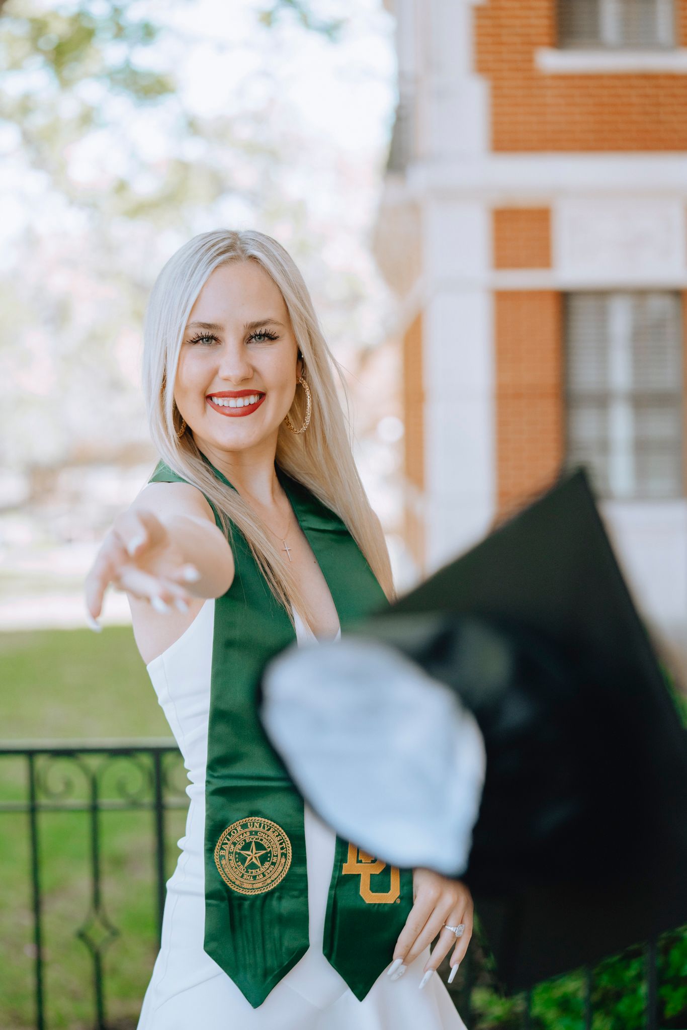 Smiling blonde woman in white dress with green Baylor University graduation stole tossing cap.