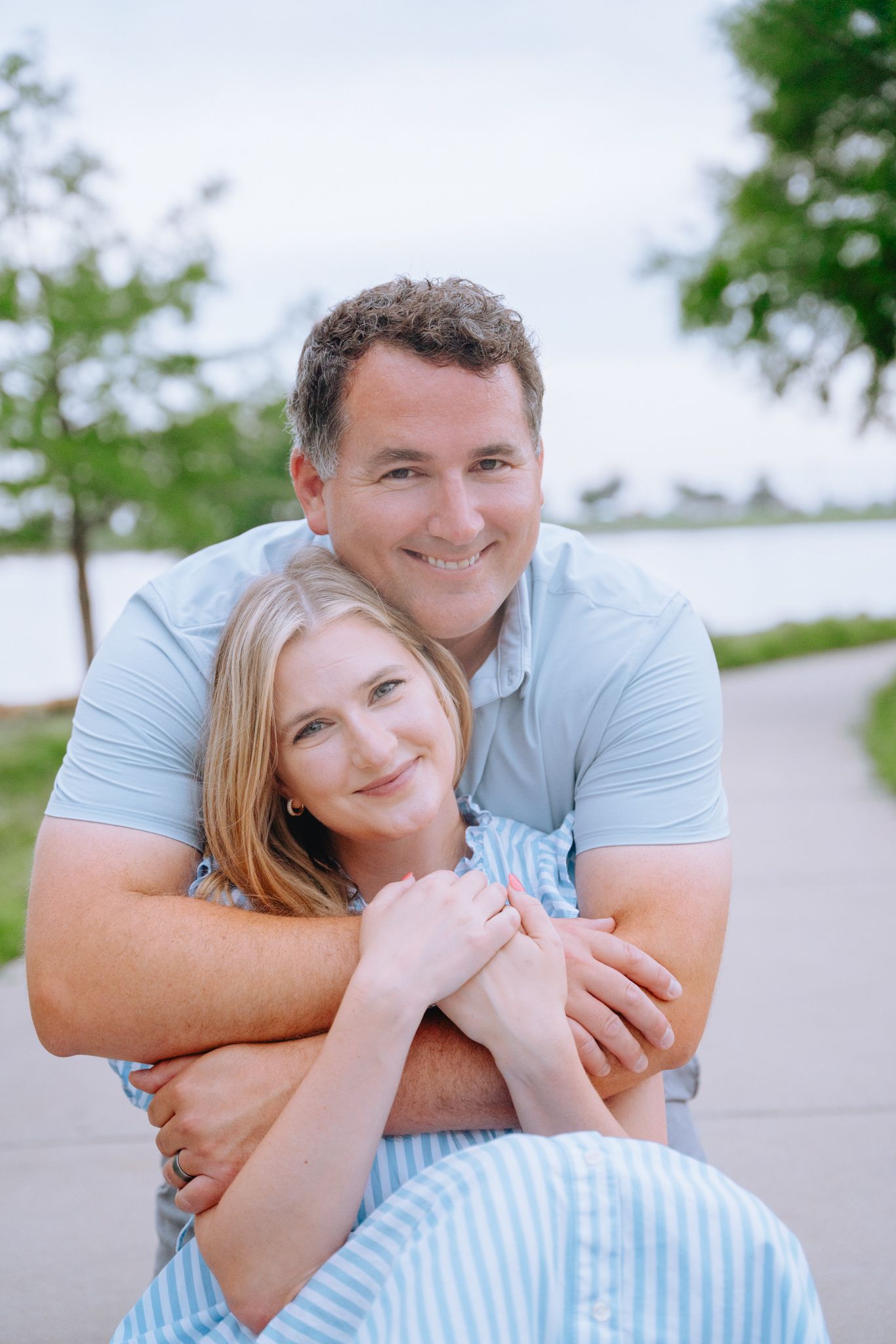 Smiling couple embracing outdoors near a lake, man in blue polo hugging woman in striped dress.