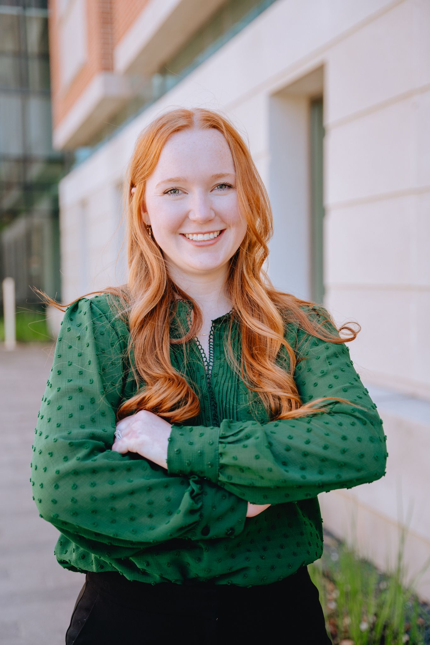 Smiling young woman with red hair in green blouse standing with arms crossed outside.