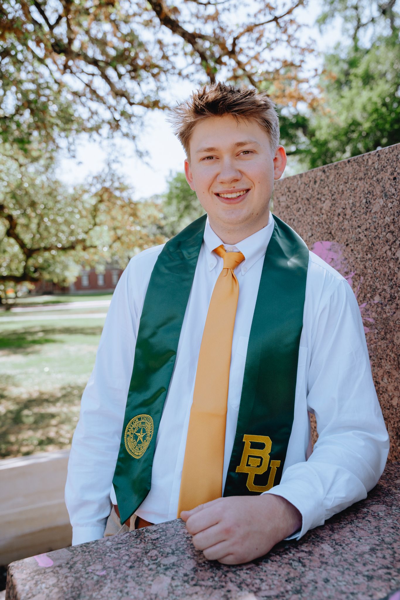Young male Baylor University graduate wearing green BU stole and gold tie smiles outdoors.