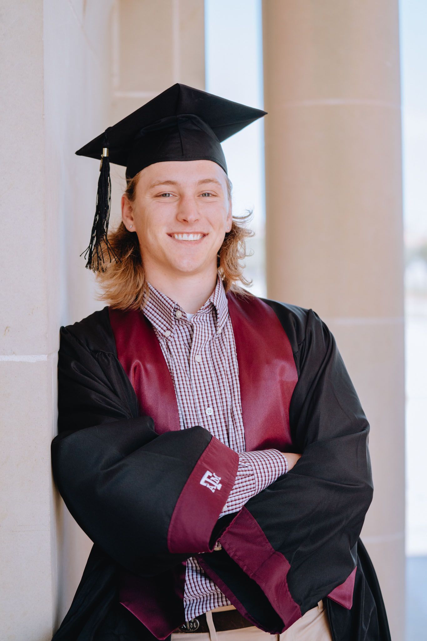 Smiling male graduate in black cap and maroon gown posing with arms crossed against white columns.