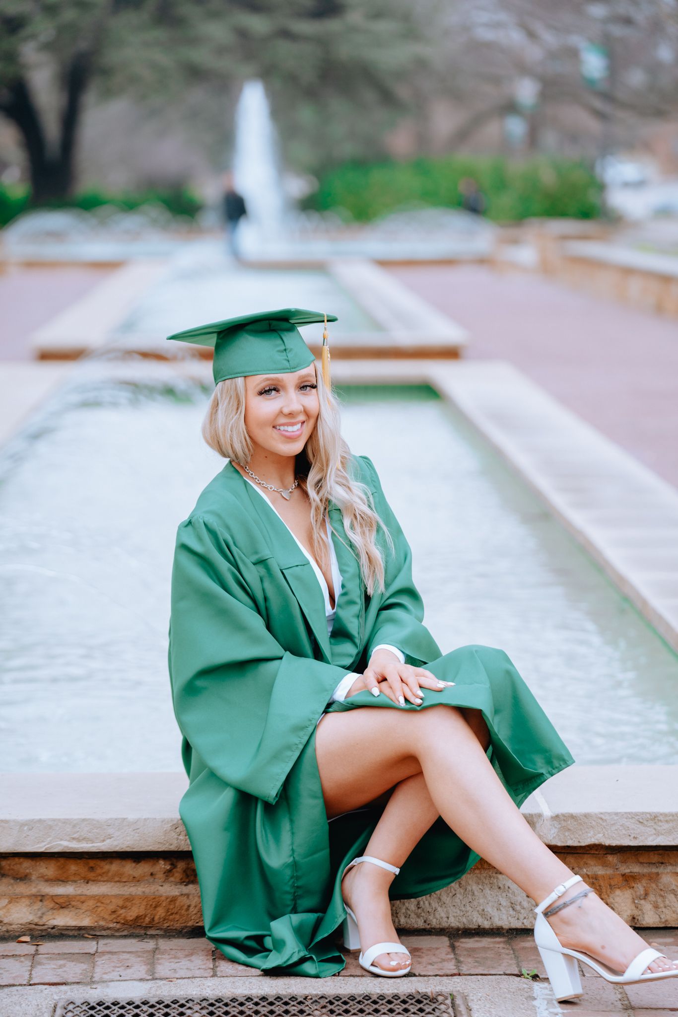 Smiling graduate in green cap and gown sitting by a fountain outdoors. florida photos