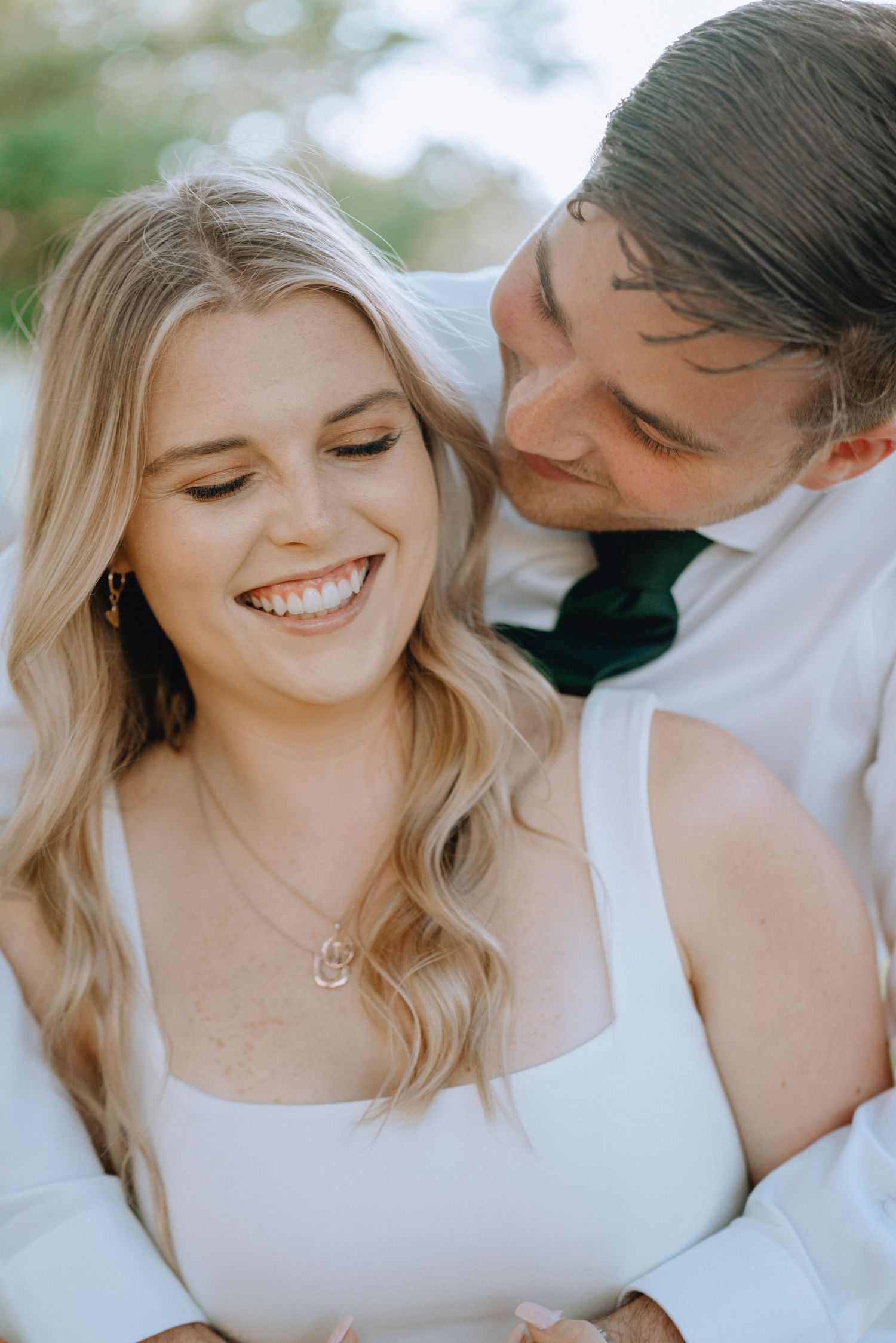Groom in white shirt and green tie embracing smiling blonde bride from behind outdoors.