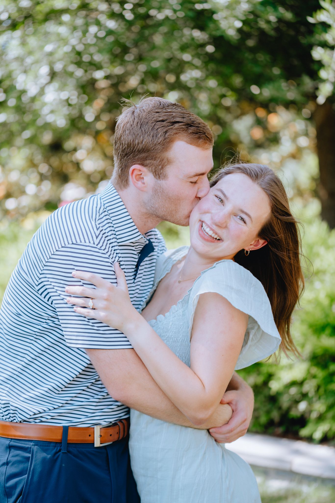 A happy couple embraces outdoors, man kissing woman on cheek as she laughs, surrounded by lush green trees.