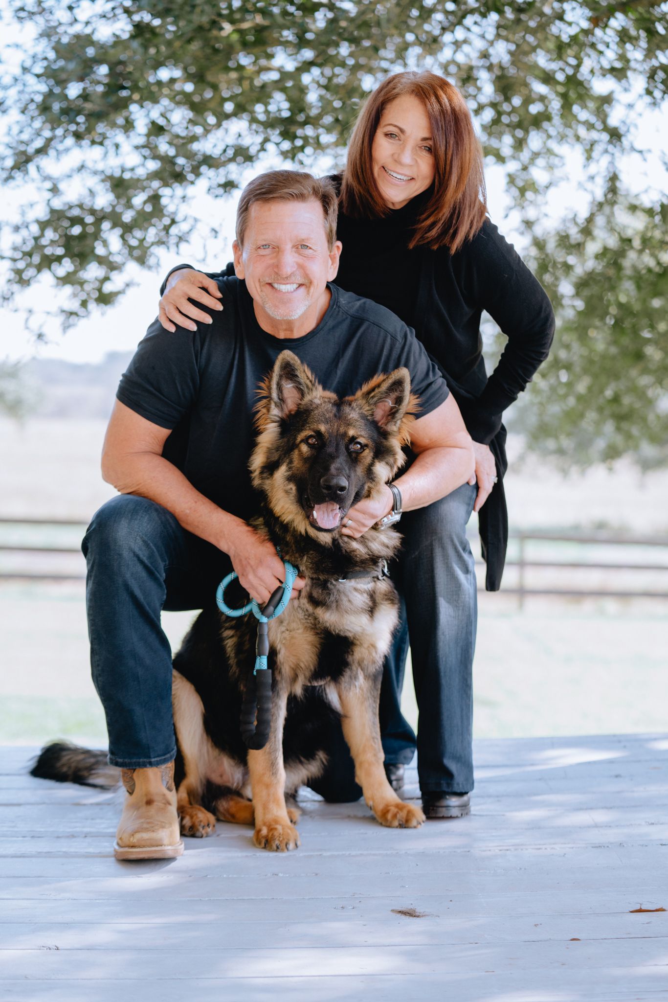 Smiling couple posing outdoors with their German Shepherd dog on a wooden deck.