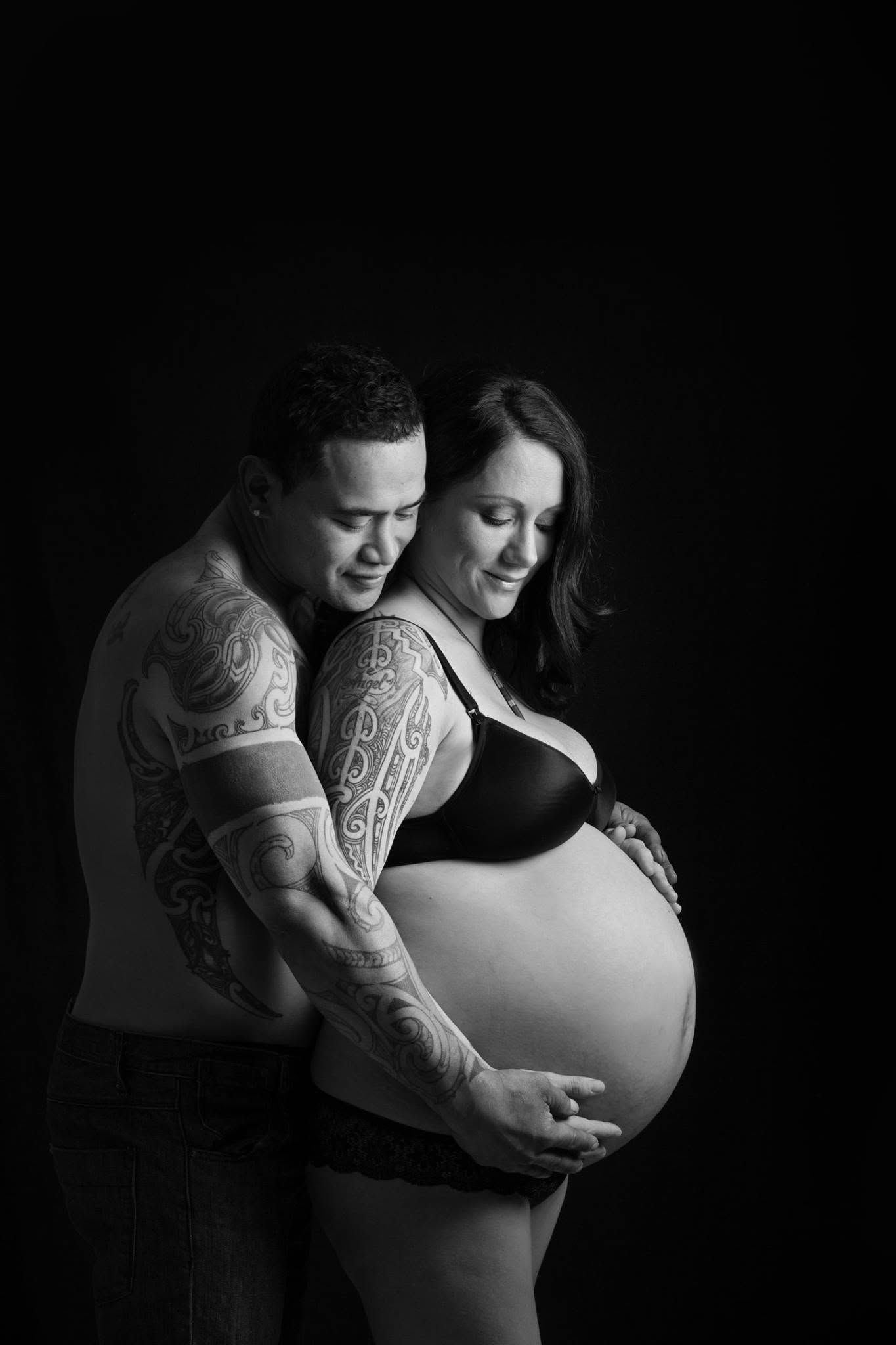 Black and white maternity photo of tattooed man embracing pregnant woman from behind.
