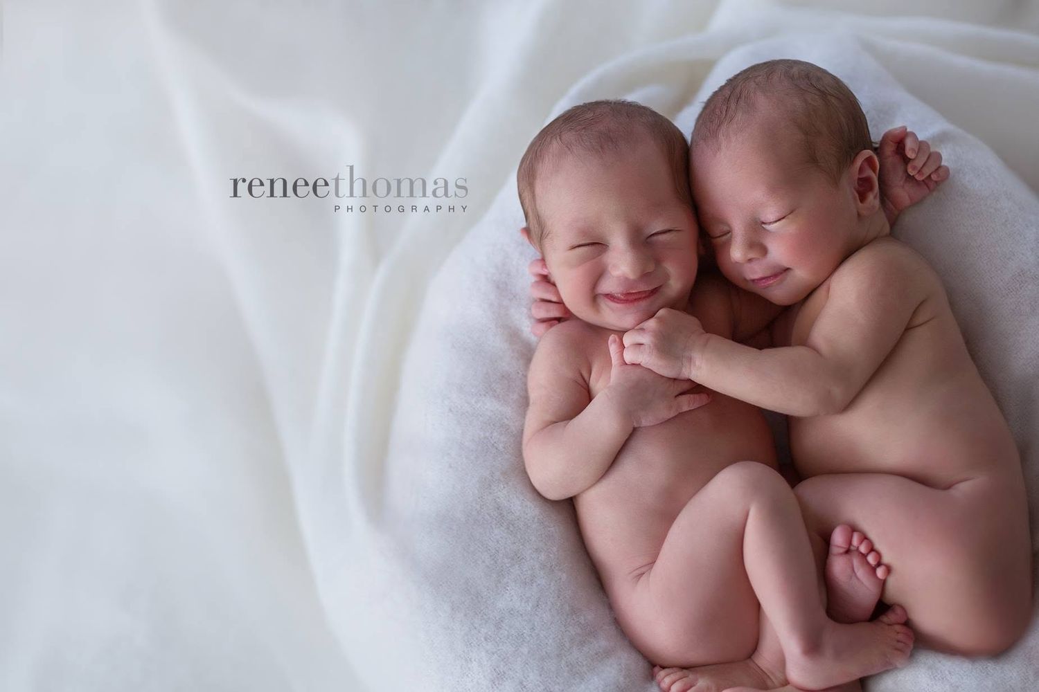 Two smiling newborn twins snuggled together on a white blanket by Renee Thomas Photography.