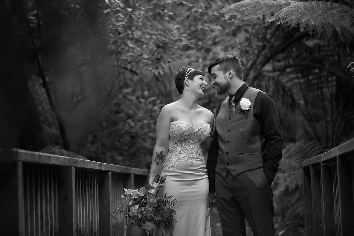 Black and white wedding portrait of bride and groom gazing at each other on wooden bridge.