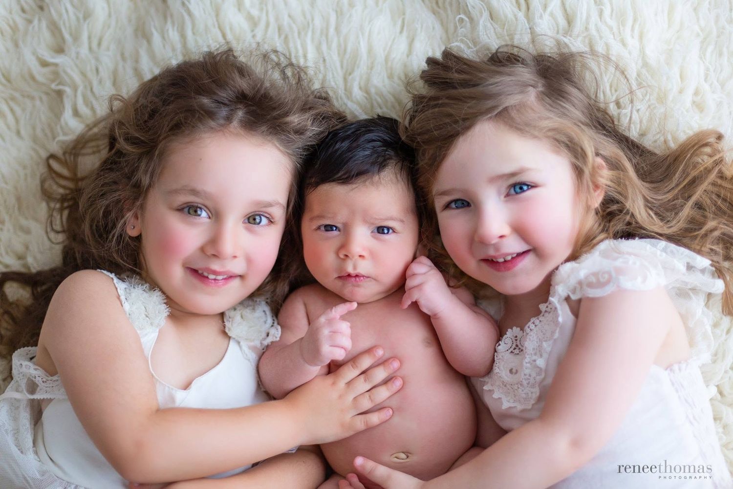 Two smiling girls in white dresses cuddle a sleeping newborn on a fluffy cream blanket.